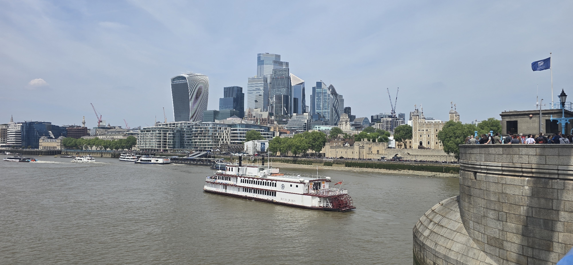This wide shot captures a cityscape of London, United Kingdom, featuring a waterway with several vessels. A prominent white steamboat with a red paddle wheel is centered in the foreground. In the background, modern skyscrapers, including the distinct "Walkie Talkie" building, rise amidst older architectural structures like the Tower of London. A stone parapet filled with people is visible on the right, alongside a flagpole bearing a blue flag. The sky is overcast, suggesting daytime under diffused light. The water is a muddy color, reflecting the ambient light.