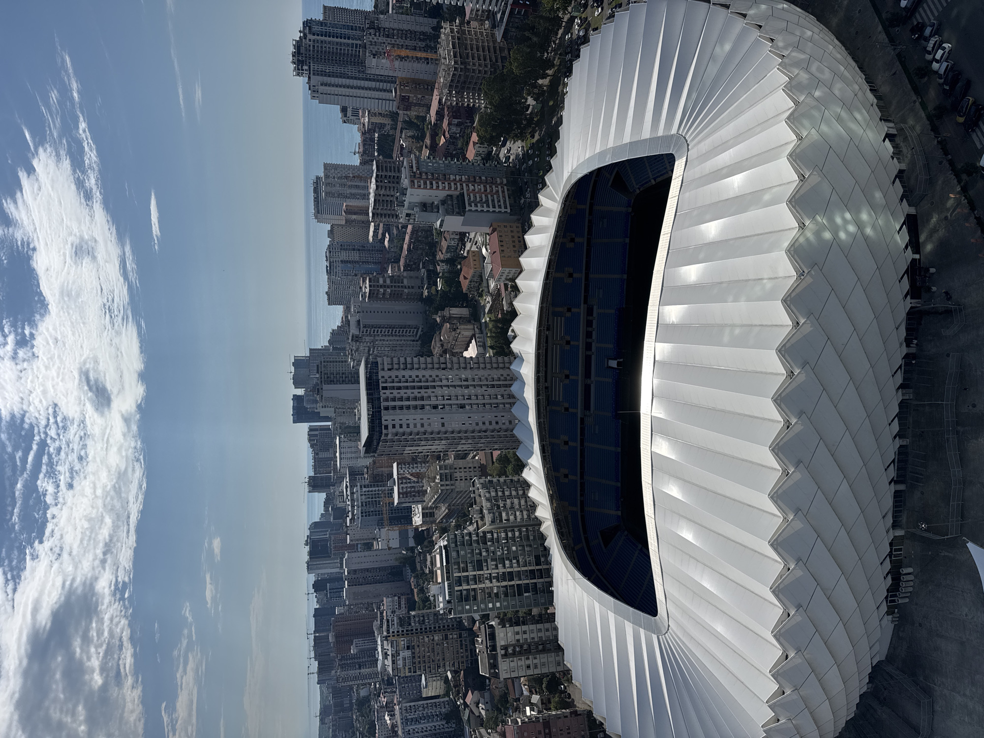 The image is an aerial view of the Batumi Stadium, a modern football stadium located in Batumi, Georgia. The stadium features a distinctive, undulating white roof designed to resemble scales or petals. The seating within the stadium is visible, appearing to be blue.
Beyond the stadium, the cityscape of Batumi unfolds. Numerous multi-story buildings, ranging in architectural style from older, block-like structures to contemporary high-rises, dominate the urban landscape. The buildings are densely packed, indicative of a coastal city. In the distance, the Black Sea stretches out, its blue waters meeting the horizon under a partly cloudy sky with visible sunbeams. The overall impression is one of a bustling coastal city with modern infrastructure juxtaposed against the natural beauty of the sea.