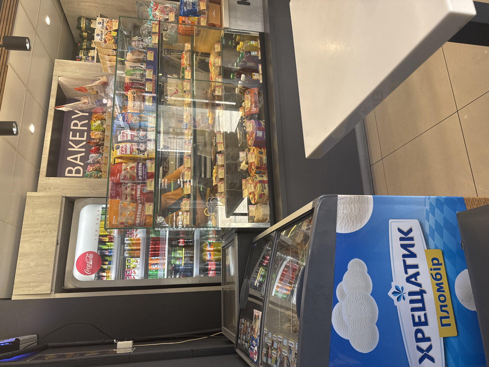 The image depicts the interior of a small food establishment, likely a convenience store or a bakery, situated in the city of Dnipro, Ukraine. In the foreground, a white table is partially visible on the right, with a tiled floor beneath it. To the left, a large blue freezer chest is prominently displayed, adorned with an advertisement for "Хрещатик Пломбір," a type of ice cream. The freezer has a depiction of clouds and ice cream scoops on its side.
Behind the freezer, a glass-doored display case showcases a variety of baked goods and packaged snacks, including cakes, pastries, and cookies. Above this display, a refrigerated Coca-Cola cooler is visible, stocked with various beverages. Further back, a prominent sign with the word "BAKERY" in large letters indicates the nature of the establishment. The shelves behind the "BAKERY" sign display more packaged food items, including coffee products. The overall environment appears to be well-lit and organized, suggesting a typical retail setting for food products.