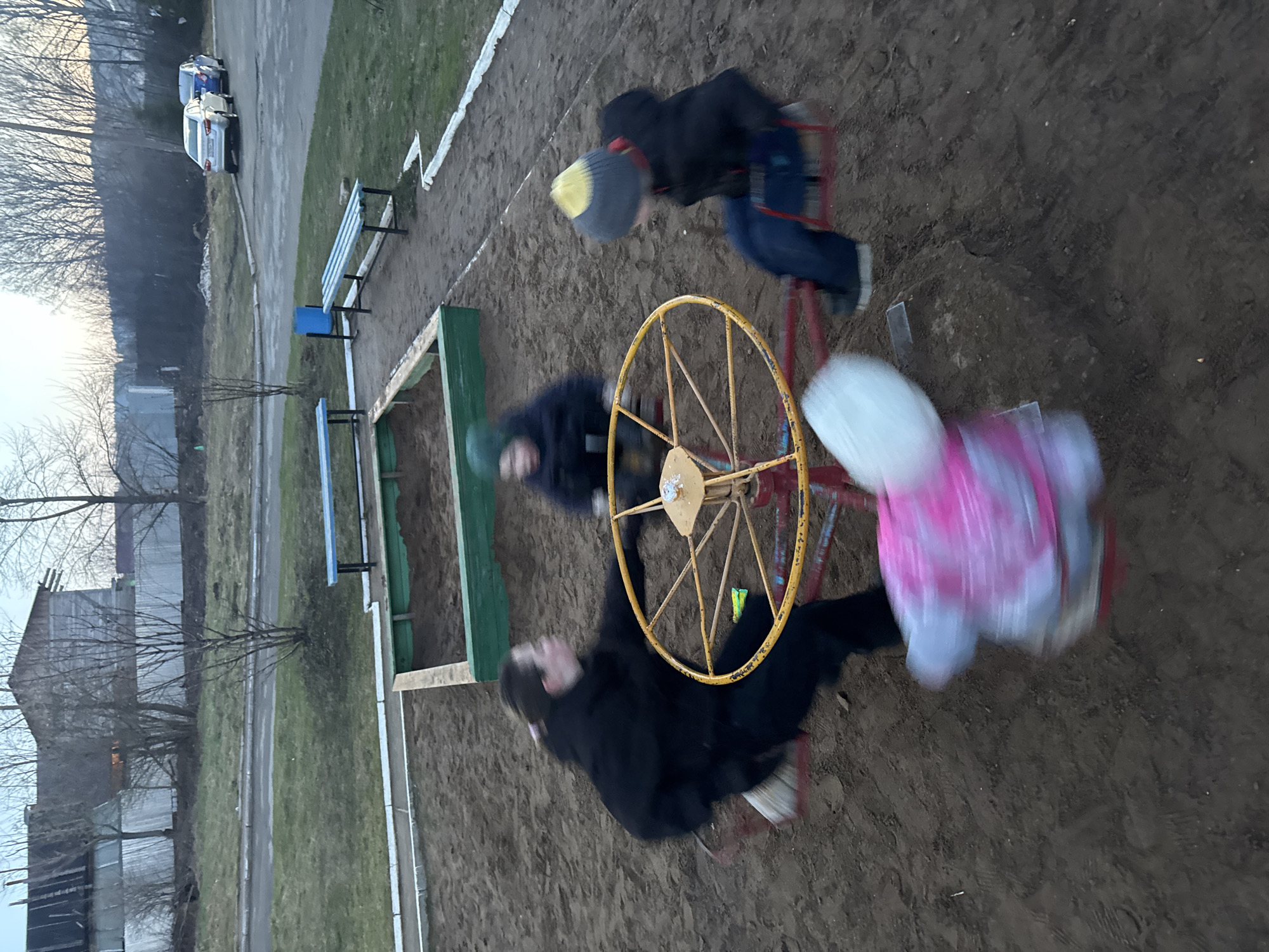 The image depicts a lively scene of children playing on a merry-go-round in a playground, likely in Chaikovsky, Russia. Four children are engaged with the yellow and red hand-powered merry-go-round, their movements blurred to convey its spinning motion. One child in a black jacket and a yellow-and-grey striped hat appears to be actively pushing the ride, while others are seated or clinging on.
The playground area consists of a dirt or sandy ground surface, with a green wooden sandpit or raised garden bed situated near the merry-go-round. Two blue-slatted benches with black frames are visible to the left, indicating a public recreation space. Beyond the immediate play area, a narrow asphalt path or road runs parallel, where a white sedan and a blue or grey vehicle are parked.
The background features numerous bare trees, suggesting either late autumn, winter, or early spring. Fences and indistinct low-rise buildings are partially visible further in the distance. The lighting is soft and diffused, indicative of an overcast sky or possibly late afternoon/early evening light, as there are no harsh shadows. The children are dressed in warm clothing, including jackets and hats, suggesting cool weather. The overall atmosphere is one of energetic outdoor play. No visible text is discernible in the image.