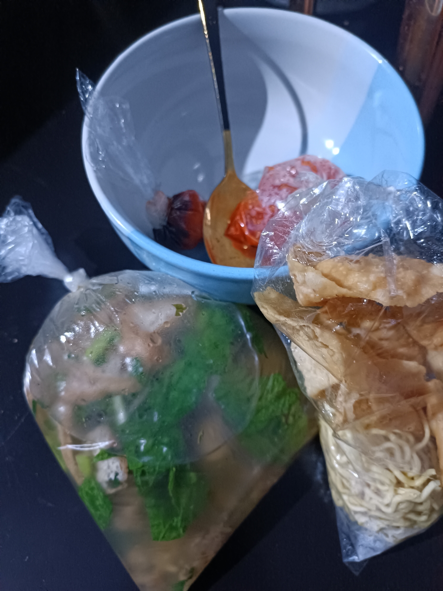 In Jetis, Indonesia, a dark surface, possibly a table, displays a collection of food items. A light blue and white bowl contains a golden spoon, which is partially submerged in what appears to be broth. Inside the bowl, small portions of red and dark brown food items are visible. To the left of the bowl, a clear plastic bag is tied shut, filled with a light-colored liquid and green leafy vegetables, with hints of cubed tofu and meat. To the right of the bowl, another clear plastic bag is tied shut, containing dry, golden-brown fried wonton wrappers and a portion of yellow noodles. The scene is dimly lit, with the focus on the food items.