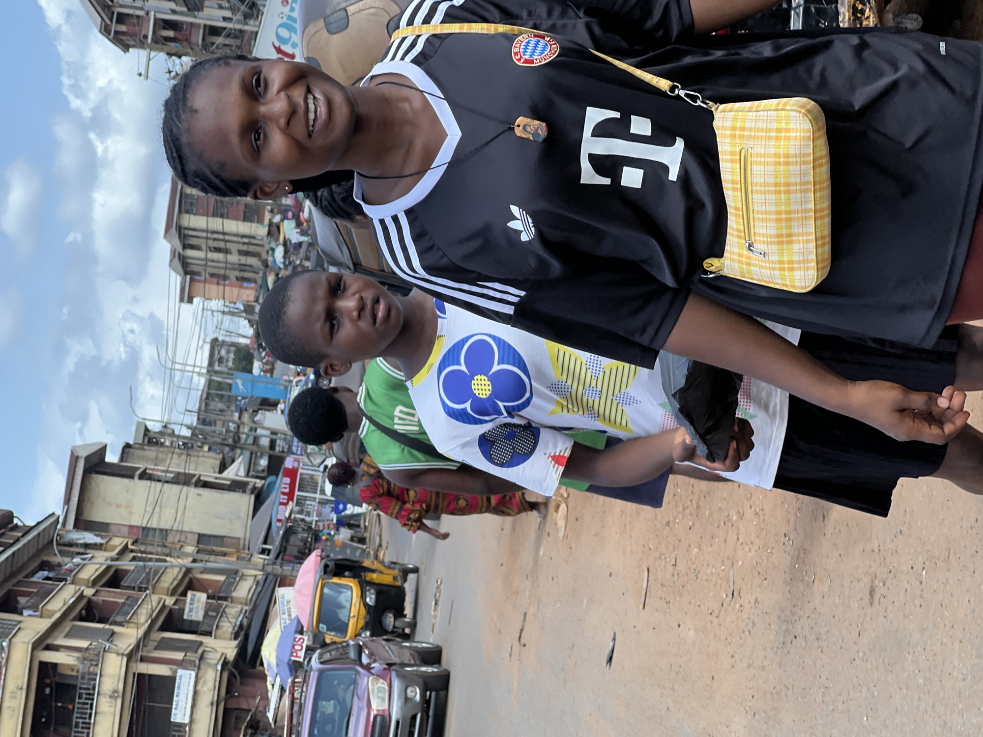 A slightly rotated full outdoor shot shows two people walking on a street.
The person in the front, on the left, is a woman with dark skin and dark hair and is smiling. She is wearing a black jersey with white stripes on the shoulder and a logo on the upper left side, and another logo in the center. She is wearing a small yellow purse on her right shoulder.
A younger person stands to her right and slightly behind her. They are wearing a white t-shirt with an image of a flower on the front and black shorts. Their expression is neutral.
Behind them are other people walking on the street. Cars and a motorcycle are parked on the street. Buildings are visible on the left side of the image. The sky is overcast.