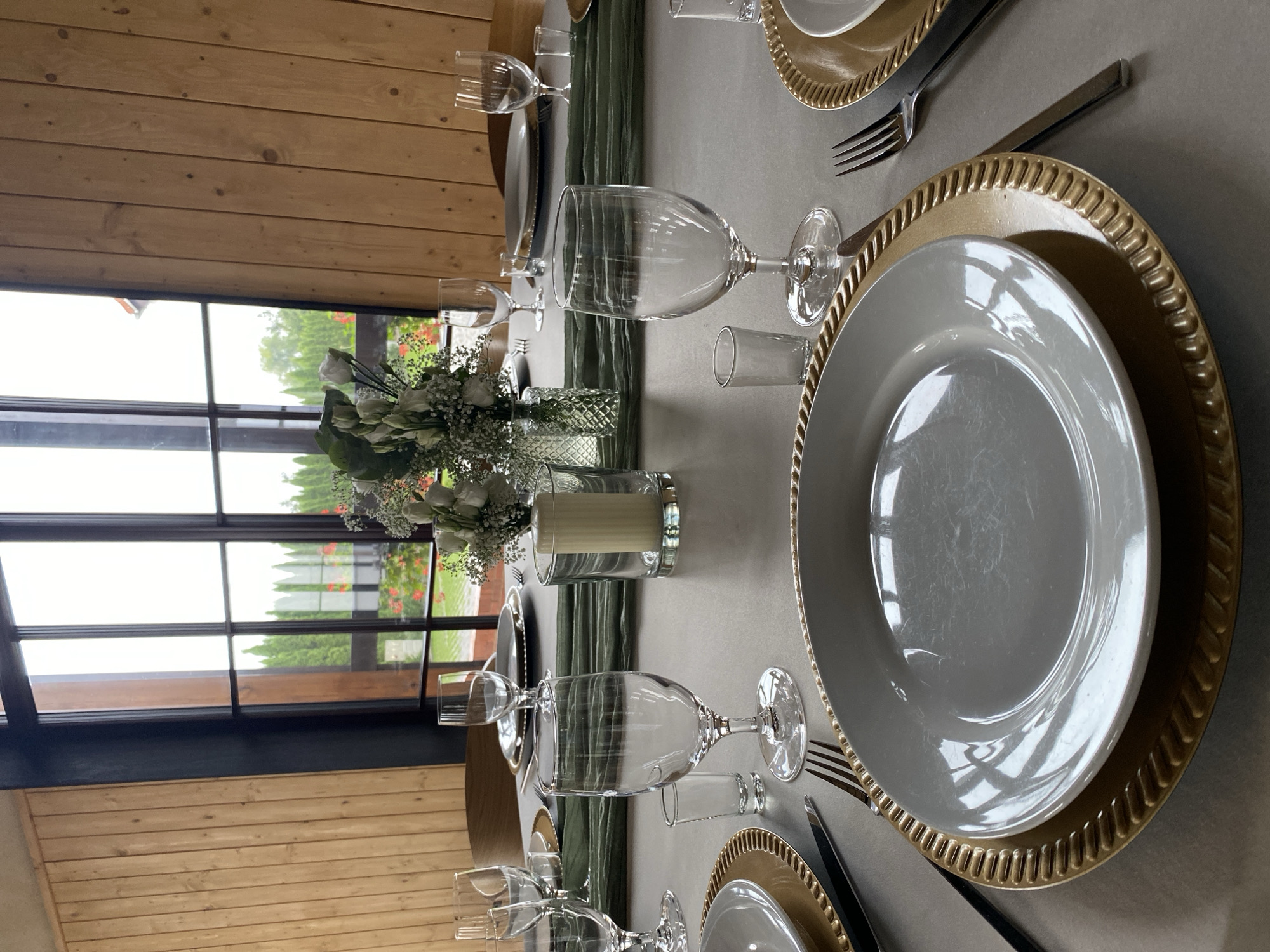 The image depicts a formally set dining table inside a room with light wood paneled walls and large windows overlooking an outdoor scene. The table is covered with a light gray tablecloth and set with white plates on gold chargers, cutlery, and glassware. A green runner drapes across the table, adorned with a floral arrangement of white roses and baby's breath in a clear vase, and a thick white candle in a glass holder. The windows reveal a view of lush green trees and foliage, suggesting a rural or park-like setting. The room's architecture features dark window frames and a wooden beam ceiling. Based on the visual cues, this scene is likely a dining area within a venue or a private residence in Buk, Poland, potentially for an event such as a wedding or a special gathering. No people are visible in the image.