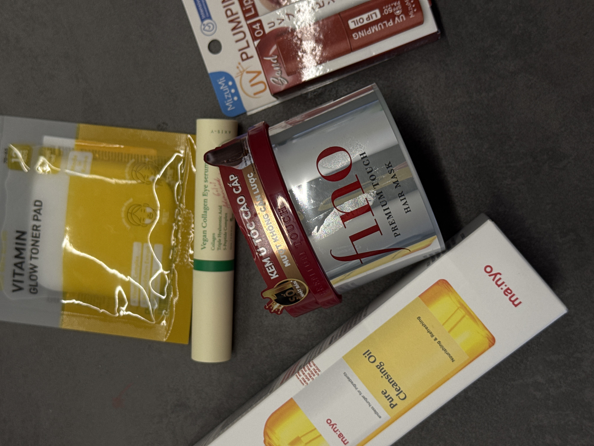 The image shows a close-up shot of several cosmetic and skincare products on a gray surface. The products include: a packet of "VITAMIN GLOW TONER PAD," a tube of "Vegan Collagen Eye serum," a jar labeled "KEM Ủ TÓC CAO CẤP" and "PREMIUM TOUCH HAIR MASK," a box of "Pure Cleansing Oil" from the brand "ma:nyo," and a package of "UV PLUMPING" lip oil from the brand "MizuMi." The image is taken from an overhead angle, showcasing the packaging and labels of the products.
