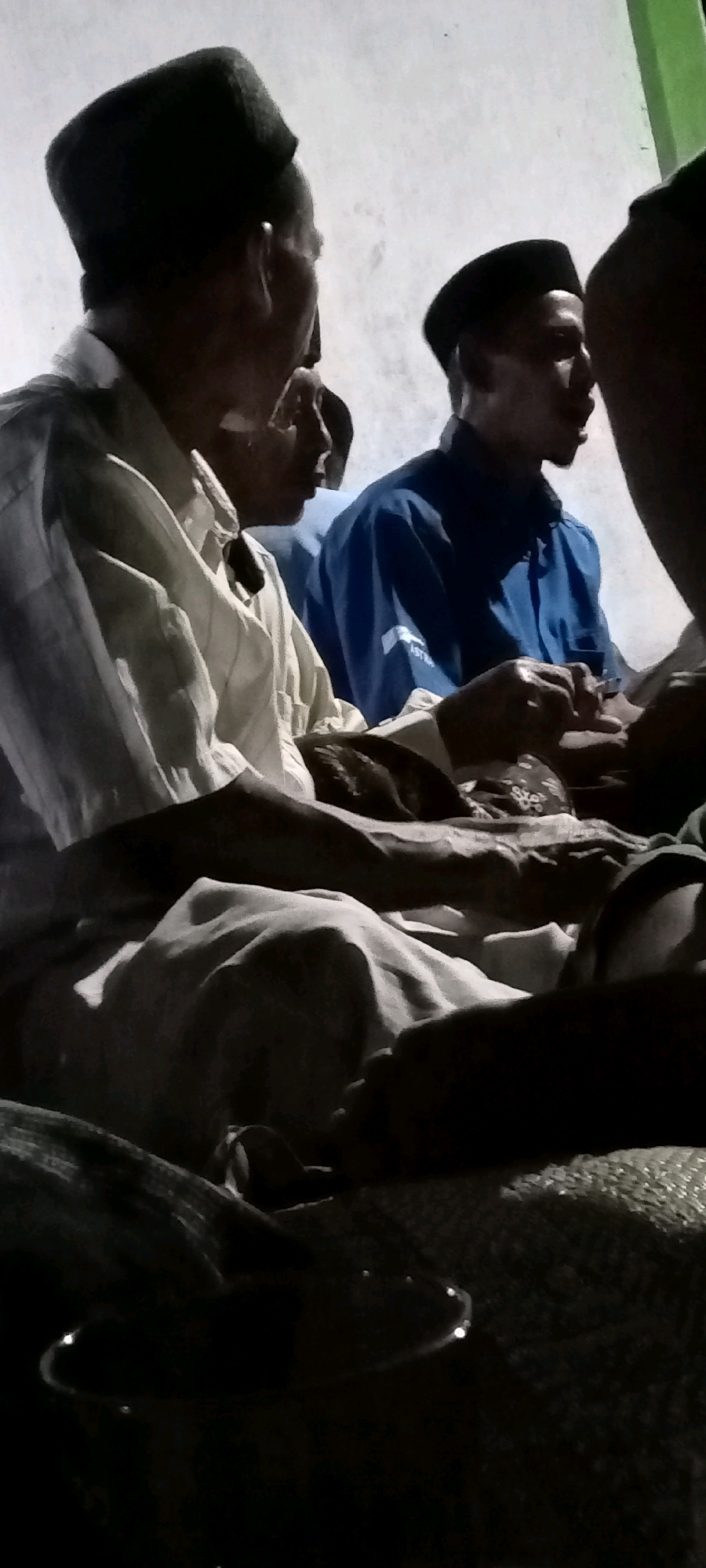 The image depicts a dimly lit scene, likely indoors, featuring at least three individuals. The foreground is dominated by the torsos and arms of two men, one in a light-colored shirt and the other in a dark blue shirt with a white logo on the sleeve. Both men are wearing black peci caps, indicating a possible religious or cultural context. Their hands are clasped or engaged in an interaction in the center of the frame. The background consists of a textured, off-white wall. The overall lighting is low, with some areas of illumination creating shadows and highlighting the textures of clothing and skin. The presence of a woven mat or carpet is suggested in the lower portion of the image, and what appear to be decorative string lights are visible in the bottom right corner. The scene appears to be a gathering or a moment of conversation. The location is identified as Kedungmulyo, Indonesia.