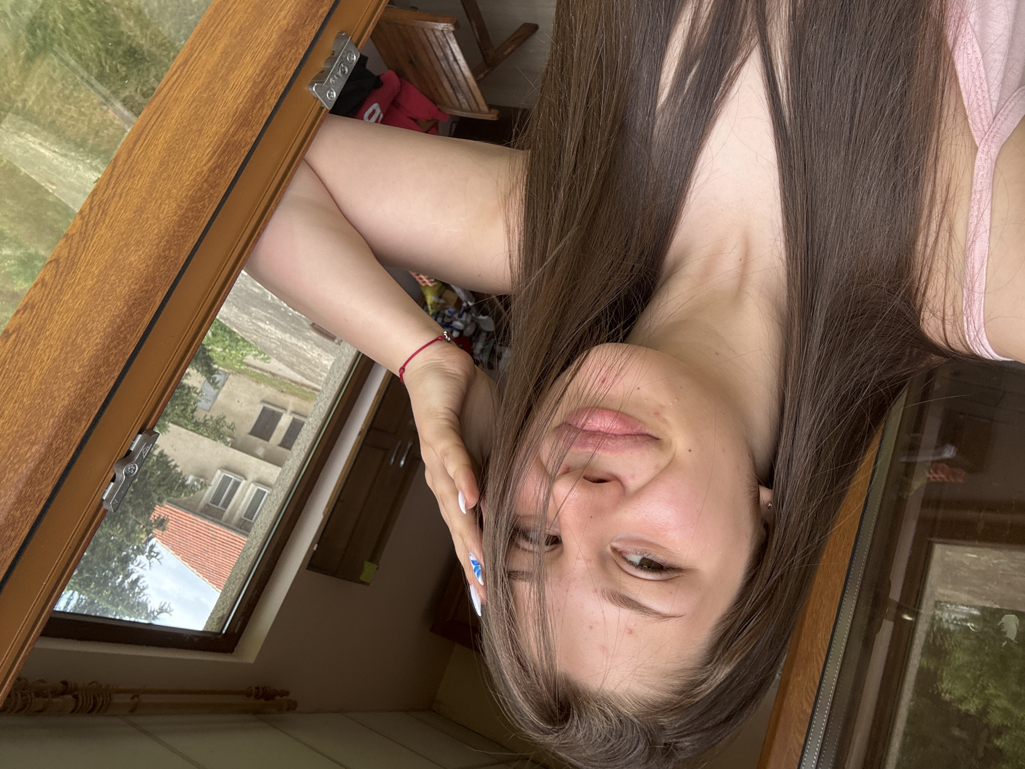 This is a portrait-oriented, upside-down, close-up photograph of a young woman with long, dark brown hair. She is positioned indoors, leaning against a wooden frame, likely a window or door. Her arms are crossed, and one hand is resting on her head, partially obscuring her face. She is wearing a light pink tank top. Her expression is relaxed and slightly playful, with a gentle smile and a direct gaze towards the camera. Her skin appears clear with a few minor blemishes. Her fingernails are painted white with blue accents.
Through the window, a view of buildings and trees is visible, suggesting an outdoor, residential setting. The time of day appears to be daytime, as natural light streams in. The sky visible through the window is a bright blue with some clouds. The overall mood of the photograph is casual and intimate. There is no visible text in the image.