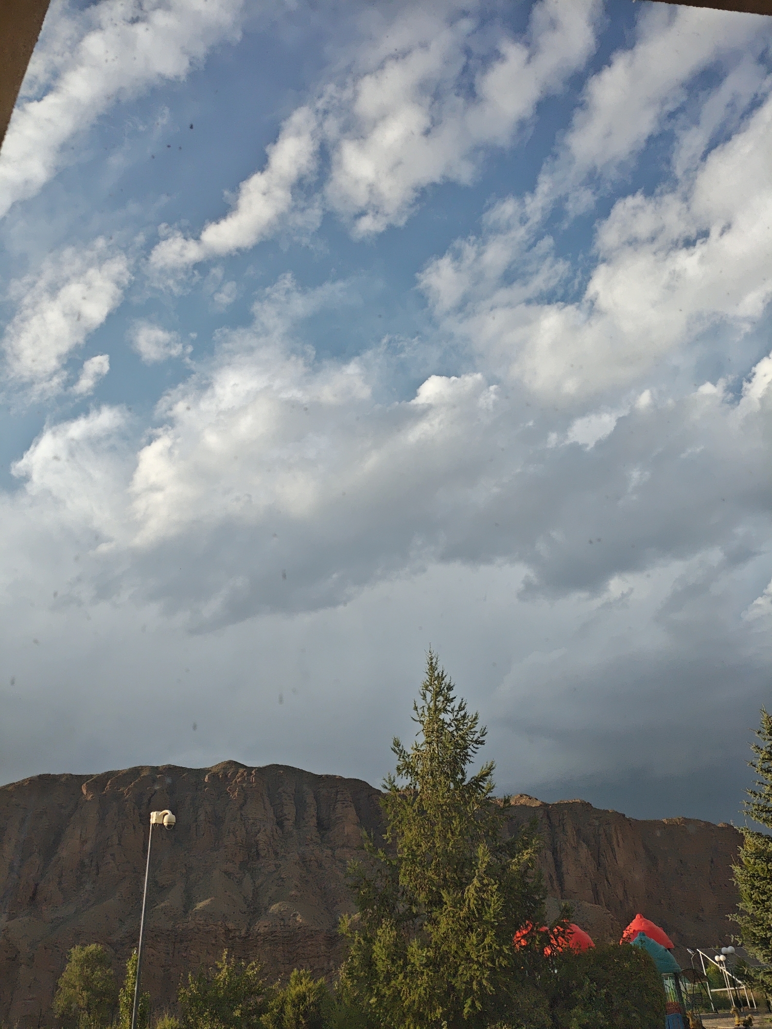 The image shows a view of a mountain range under a dramatic sky. The sky is a mix of blue and grey clouds, with some patches of white, fluffy clouds. The mountain range is large and appears to be made of reddish-brown rock, with visible striations and contours. In the foreground, there are several evergreen trees, with one particularly tall tree in the center of the frame. There are also some red and blue objects visible among the trees, which appear to be part of a playground or recreational area. A tall lamppost with a camera or surveillance device is also visible in the foreground, positioned in front of the mountain. The overall scene suggests an outdoor environment, likely a park or an area adjacent to a natural landscape. The lighting indicates it might be late afternoon or early evening, with the sun casting shadows and highlighting certain features of the mountain.