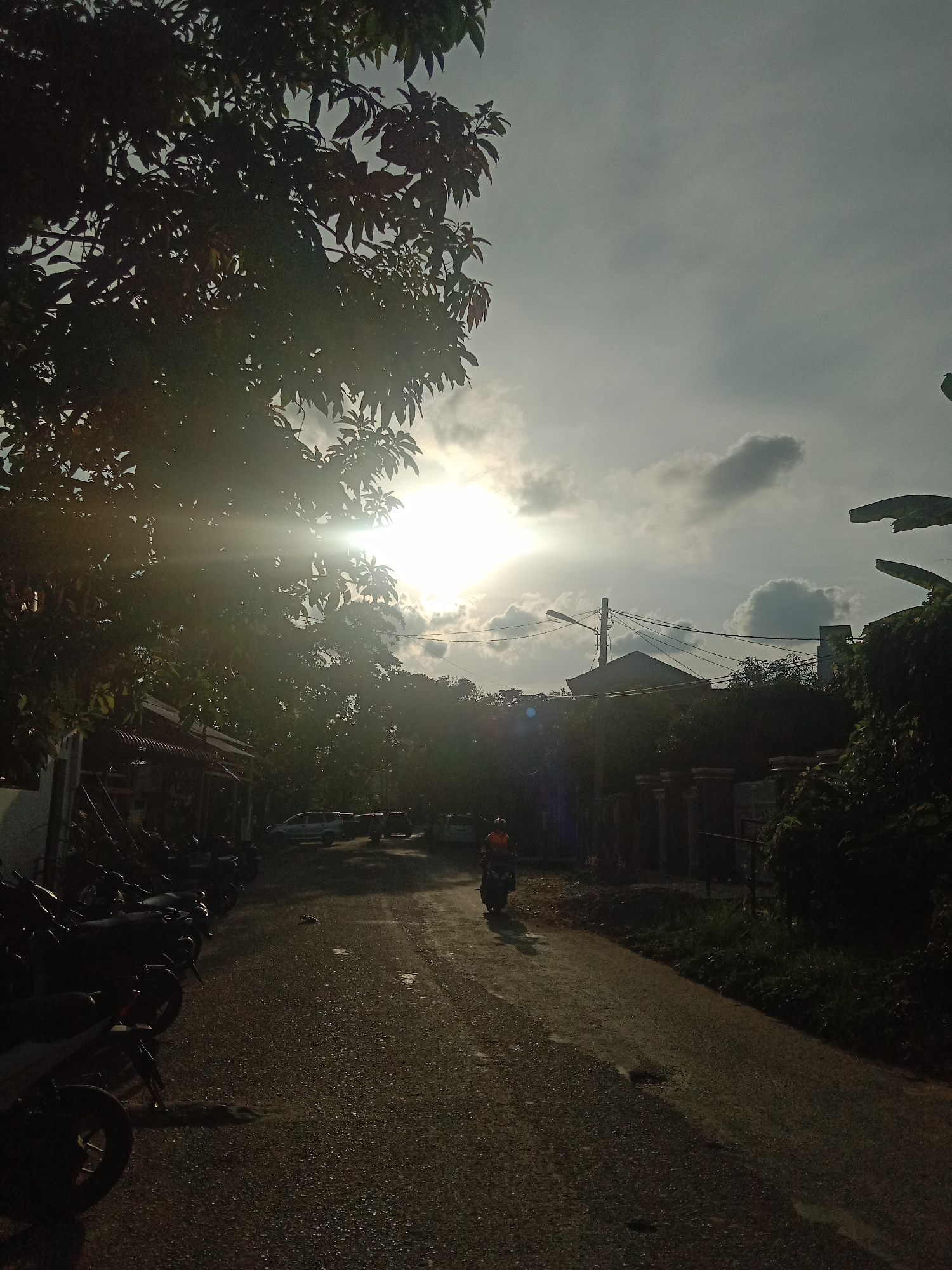 The photograph shows a sunlit street in a residential area. The sun is bright, partially obscured by clouds and trees, creating glare. The street is asphalt. A person on a motorcycle is driving away from the camera. Parked motorcycles are visible on the left side of the street, along with buildings. Trees and vegetation line both sides of the road. Overhead power lines and a street lamp are in the background. The location is identified as Lhokseumawe, Indonesia.