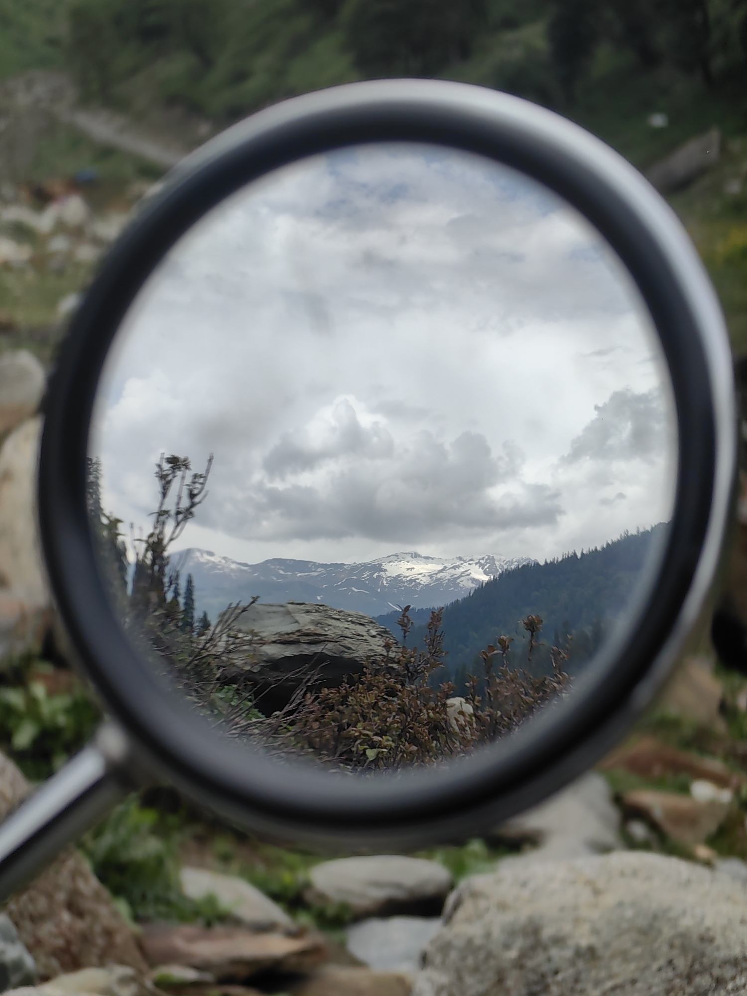 This is a medium shot, eye-level, natural-light photograph taken in Dhundi, India. The image features a close-up view through a magnifying glass, which acts as a circular frame, revealing a landscape scene beyond. The magnifying glass itself is a prominent object in the foreground, with its metallic handle visible on the left side of the frame. The handle appears to be made of polished metal.
The landscape viewed through the magnifying glass depicts a mountainous region under a cloudy sky. In the mid-ground, there are dark green coniferous trees forming a dense forest on the slopes of hills. Above the treeline, snow-capped mountain peaks are visible, suggesting a high-altitude location. The sky is filled with dramatic, heavy grey clouds, indicating overcast or potentially stormy weather.
In the immediate foreground, as seen through the magnifying glass, there are some dry, leafless branches and twigs, possibly from a bush or small tree, positioned in front of the mountain view. There are also a few large, grey rocks within the magnified scene. The area outside the magnifying glass is blurred and out of focus, showing more rocks and green vegetation, suggesting the surrounding environment is a natural, possibly rocky, terrain with some greenery.
The overall impression is one of a natural landscape being examined or magnified, drawing attention to the details of the distant mountains and the immediate foreground elements within the frame of the magnifying glass. There are no people or animals visible in the photograph.