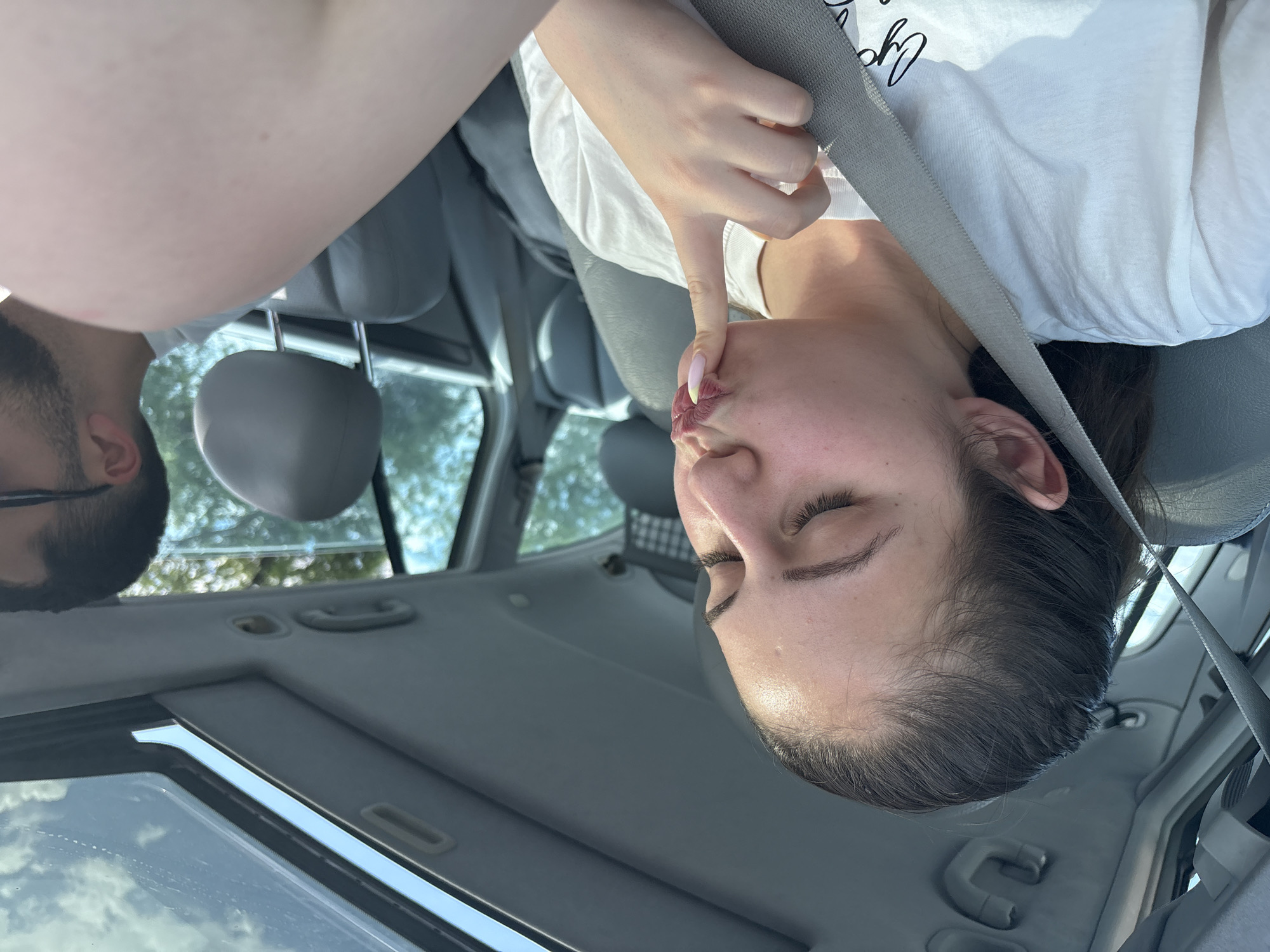 A woman is sitting in the passenger seat of a car, wearing a white t-shirt and a seatbelt. Her eyes are closed, and she's touching her lips with her index finger. A man with a beard and glasses is in the driver's seat. The location appears to be inside the car, with the interior and exterior visible through the windows. It is not possible to determine the city of Burgas, Bulgaria from the image.