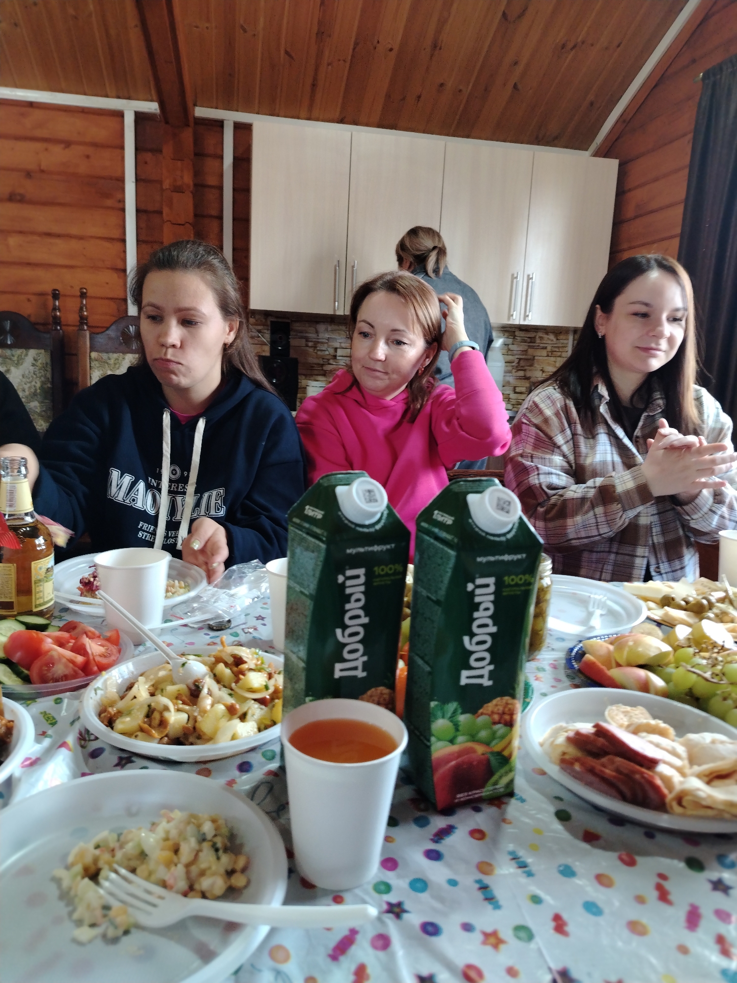 The image captures an indoor scene where three women are seated at a table laden with food and drinks. The setting appears to be a wooden cabin with wood paneling on the walls and ceiling.
The woman on the left is wearing a dark blue hoodie with white lettering and is looking down at her plate. The woman in the center is wearing a bright pink hoodie and has her hand raised to her hair. The woman on the right is wearing a brown plaid shirt and has her hands together, looking off to the side.
The table is covered with a colorful patterned plastic tablecloth and holds various dishes. Visible are salads, sliced tomatoes and cucumbers, a platter of fruit and cheese, and a plate with what appears to be sliced meat and flatbread. There are also two cartons of juice with the brand name "Добрый" (Dobryy) in Russian, along with a white cup filled with juice. An open bottle of beer is visible near the woman in the blue hoodie.
In the background, there is a person standing near light-colored cabinets. A brown curtain is also visible on the right side of the image.