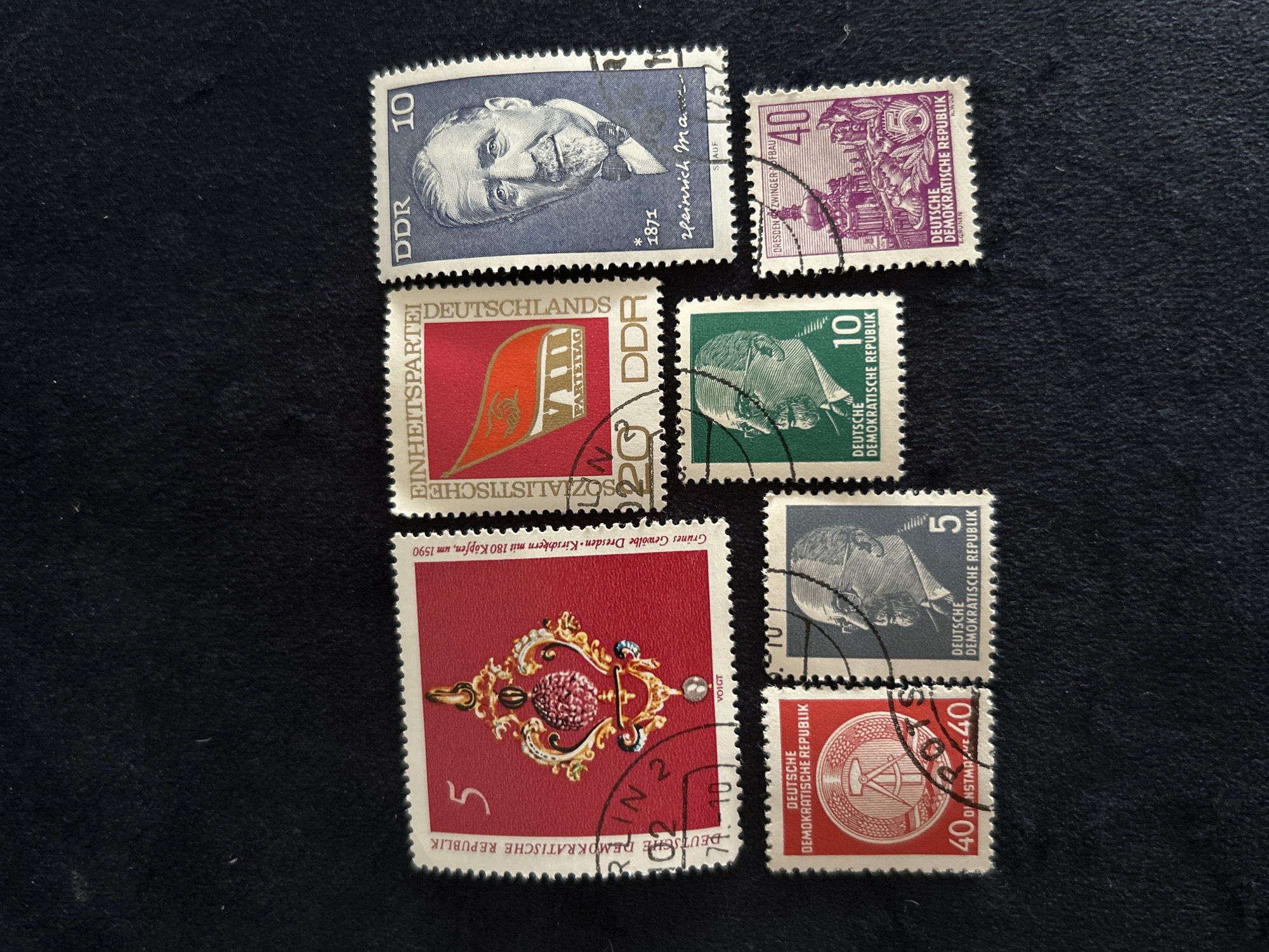 The image displays a collection of seven postage stamps arranged on a dark, textured surface. The stamps are from the German Democratic Republic (DDR) and depict various designs.
From left to right, top row:
1. A red stamp with a gold and red pendant design, featuring the number "5" and "DEUTSCHE DEMOKRATISCHE REPUBLIK".
2. A red stamp with an orange flag and the text "EINHEITSPARTEI" and "VIII PARTEITAG DDR", with the number "20".
3. A blue stamp with a portrait of a man, the text "DDR", the number "10", and "1871 heinrich man."
From left to right, bottom row:
4. A red stamp with a circular emblem featuring a hammer and sickle, the text "DEUTSCHE DEMOKRATISCHE REPUBLIK", and "40".
5. A blue-grey stamp with a profile portrait of a man with glasses, the text "DEUTSCHE DEMOKRATISCHE REPUBLIK", and the number "5".
6. A green stamp with a profile portrait of a man with glasses and a beard, the text "DEUTSCHE DEMOKRATISCHE REPUBLIK", and the number "10".
7. A purple stamp with an architectural building, the text "DEUTSCHE DEMOKRATISCHE REPUBLIK", and the number "40".
The stamps appear to be in good condition, with clear imagery and perforations.