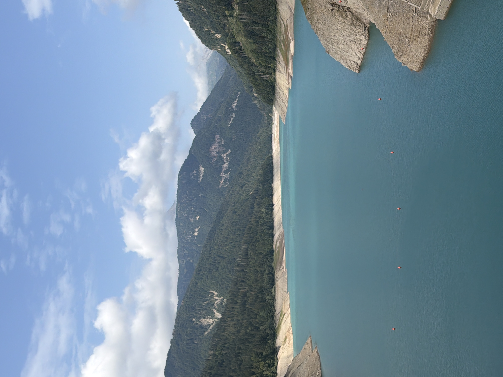 The image depicts a wide, turquoise lake surrounded by lush, tree-covered mountains. The sky above is a bright blue with scattered white clouds. In the foreground, the calm water stretches towards the distant mountains. Several small, orange buoys are visible on the surface of the lake, marking a section of it. The shoreline is characterized by rocky, eroded slopes, with some exposed dry land indicating a lower water level. On the right side of the image, a section of a concrete structure is visible along the bank. The overall scene is a serene natural landscape. The location is the Ampezzo/Dimpeç area in Italy.