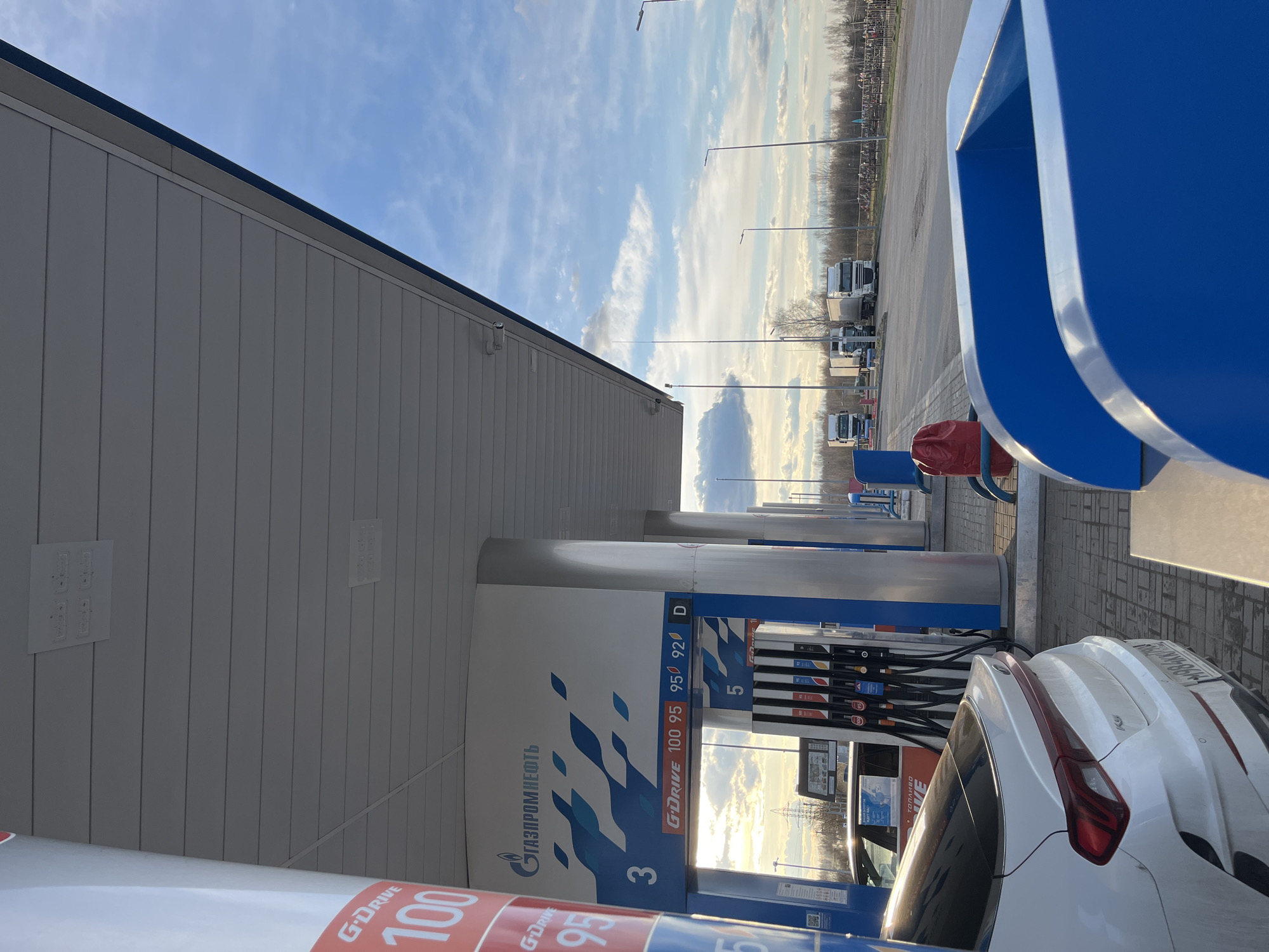 The image shows a gas station in Klin, Russia. The station features the branding of Gazprom Neft, offering G-Drive 100, 95, and 92 octane fuel, and diesel ("D"). A white car is parked at one of the pumps, with its rear end visible in the foreground. The fuel pumps are numbered, and the price display screens are visible. In the background, beyond the gas station canopy, are parked trucks, light poles, and the sky with scattered clouds.