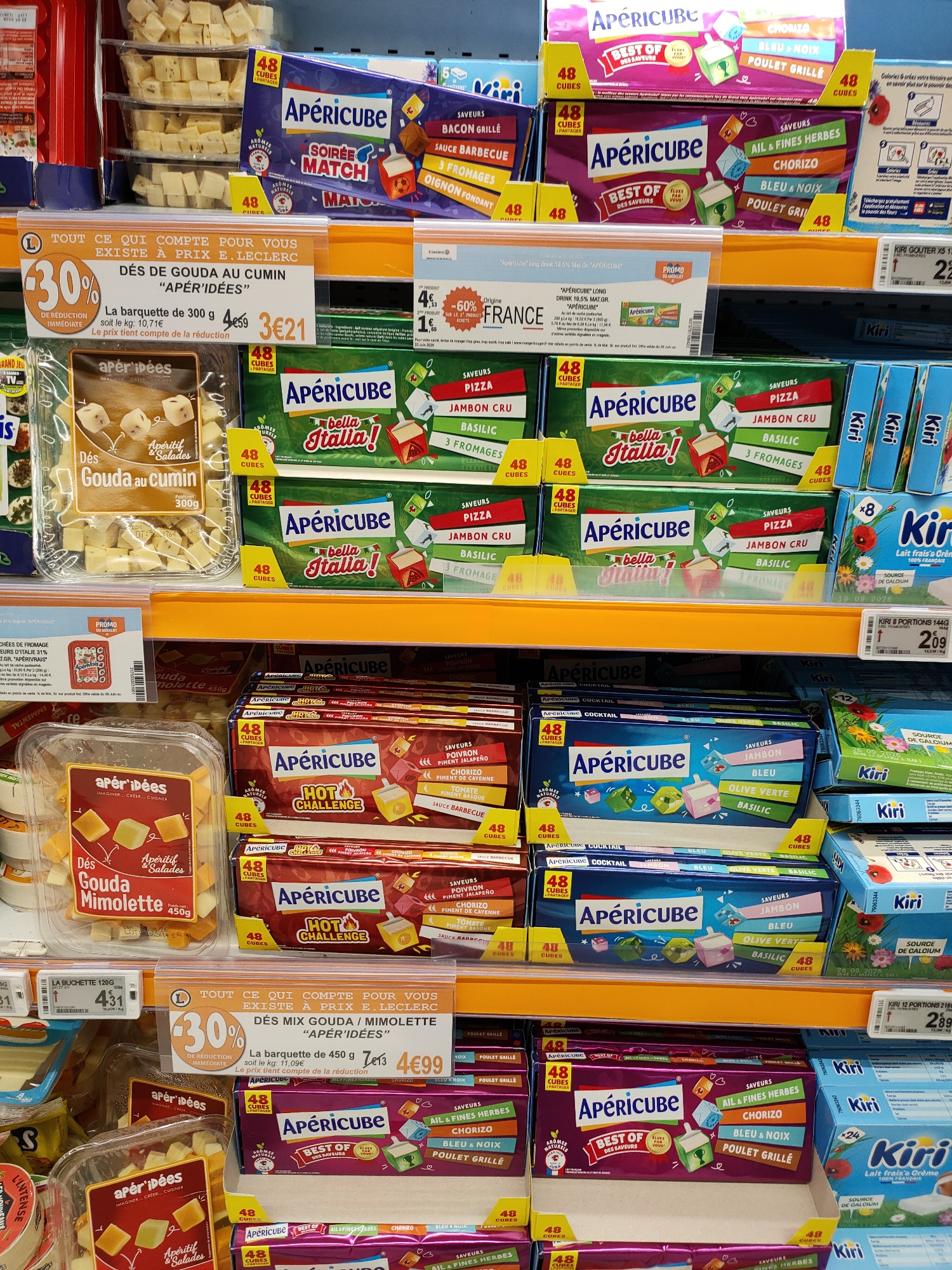 The image shows a close-up, eye-level view of a supermarket aisle stocked with various cheese products. The lighting is bright, typical of a retail environment.
In the foreground, on a lower shelf, several clear plastic containers filled with diced cheese are visible. One container, labeled "Apér'idées," contains cubed Gouda with cumin. Next to it is another "Apér'idées" container with cubed cheese labeled "Gouda Mimbolette". Further to the right are various brightly colored boxes of Apéricube cheese snacks.
The middle shelf displays more boxes of Apéricube, featuring different flavor combinations like "Bella Italia" and "Hot Challenge." There are also packages of Kiri cheese, particularly "Kiri Lait frais's Crème."
The top shelf holds more containers of diced cheese, also seemingly from the "Apér'idées" brand.
Promotional signs are present, with one prominent orange sign advertising "30% DE REDUCTION IMMEDIATE" on "DÉS MIX GOUDA / MIMOLETTE 'APÉR'IDÉES'." Another sign indicates a price of "4€99" for a "barquette" of 450g.
The products are arranged neatly on the shelves, suggesting an organized retail display. The overall scene depicts a typical grocery store cheese section. There are no people or other distinguishing environmental features visible beyond the products and shelving.