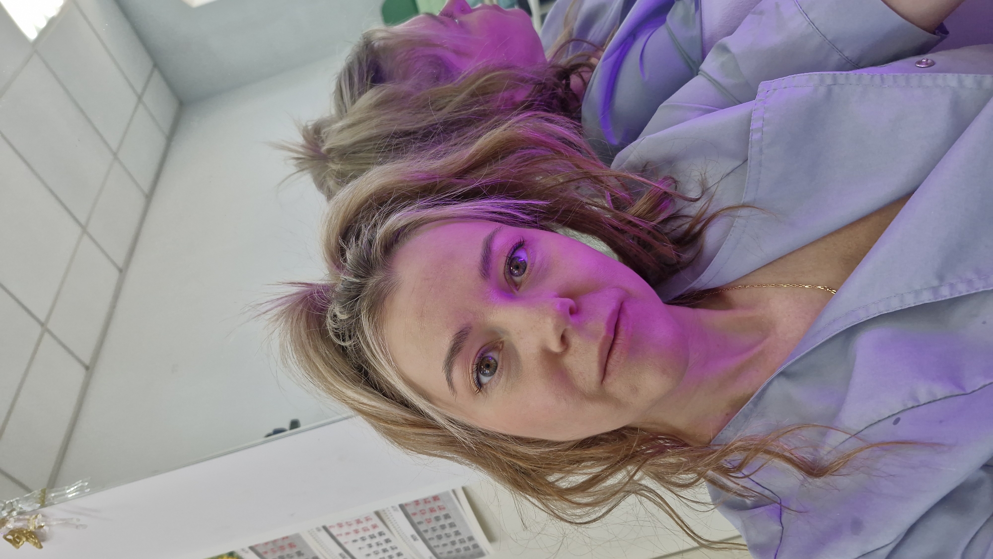A fair-skinned woman with blonde hair is looking directly at the camera in a close-up shot. She is wearing a light blue button-down shirt and a delicate gold necklace. Her reflection is visible in a mirror behind her. In the background, partially obscured by the mirror's edge, are some papers with text and possibly numerals. The lighting is predominantly pink. There is no information to determine the location in the city of Otchor, Russia from the image.