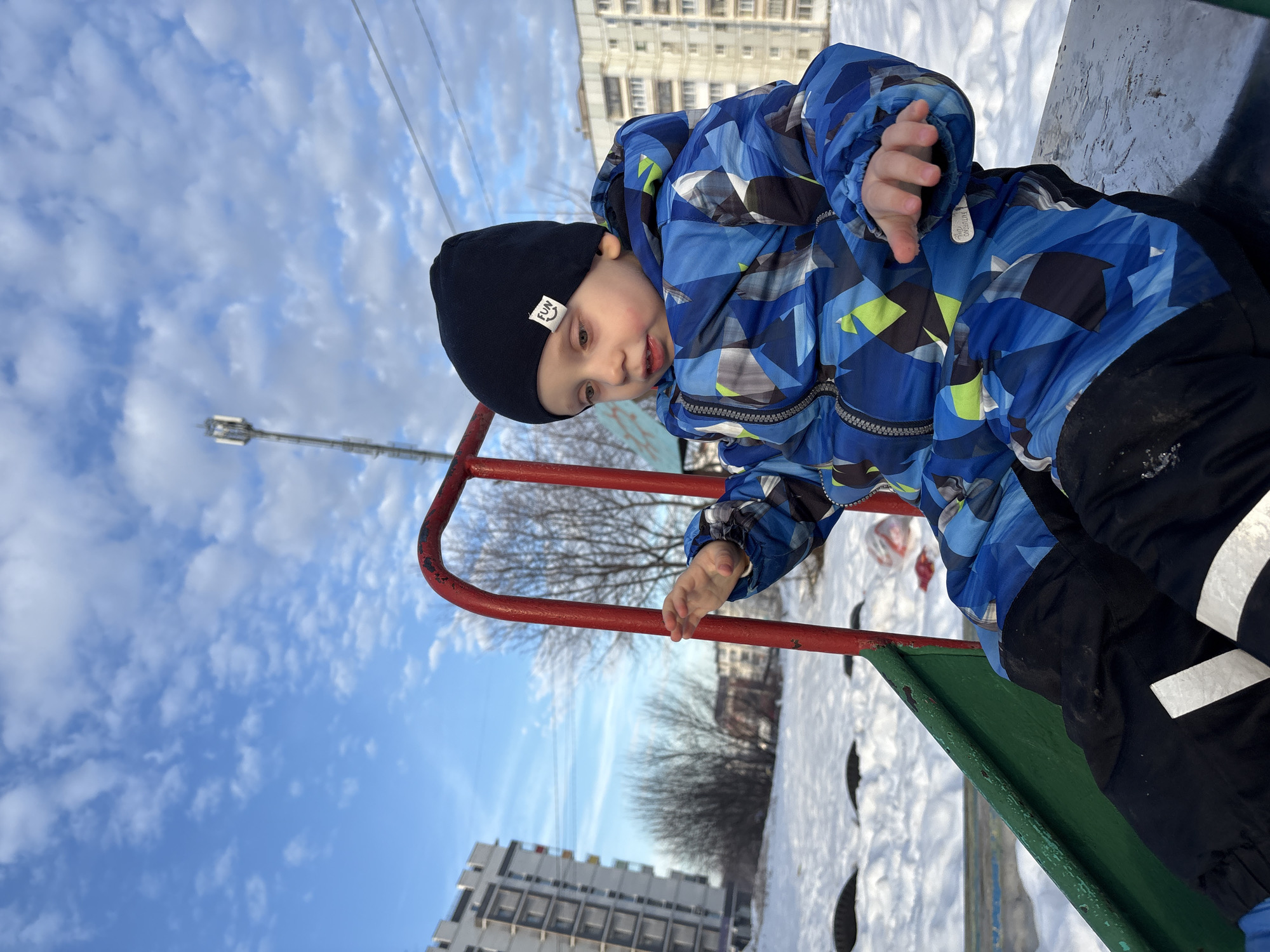 A young child, clad in a black knit beanie with a white "FUN" label, a blue, black, and lime green geometric-patterned winter jacket, and black snow pants, is seated on a playground slide in Chaikovsky, Russia. The child's hands are slightly raised with open palms. The slide structure features red metal railings and a green chute segment. The surrounding ground is blanketed in snow. In the background, several multi-story residential apartment buildings are visible, alongside bare deciduous trees. A tall communications tower stands in the distance. The sky is blue with scattered white clouds, and utility lines are discernible overhead.