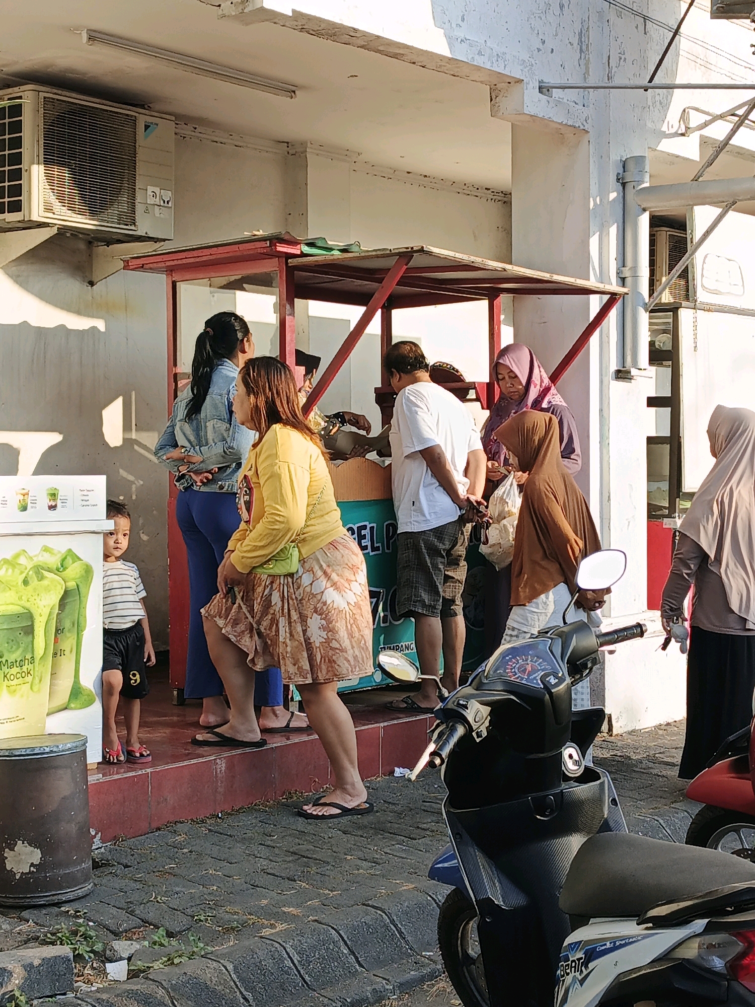 In Boro, Indonesia, a street scene unfolds in front of a vendor's stall. Several individuals, including women in hijabs and a man in a white t-shirt and plaid shorts, are gathered around the stall. A young boy in a striped t-shirt and black shorts stands near a display of green beverages. The stall itself has a red frame and is topped with a white awning. In the foreground, two motorcycles are parked, one a blue and white scooter and the other a red scooter partially visible. The ground is paved with interlocking grey stones. An air conditioning unit is mounted high on the wall above the stall.