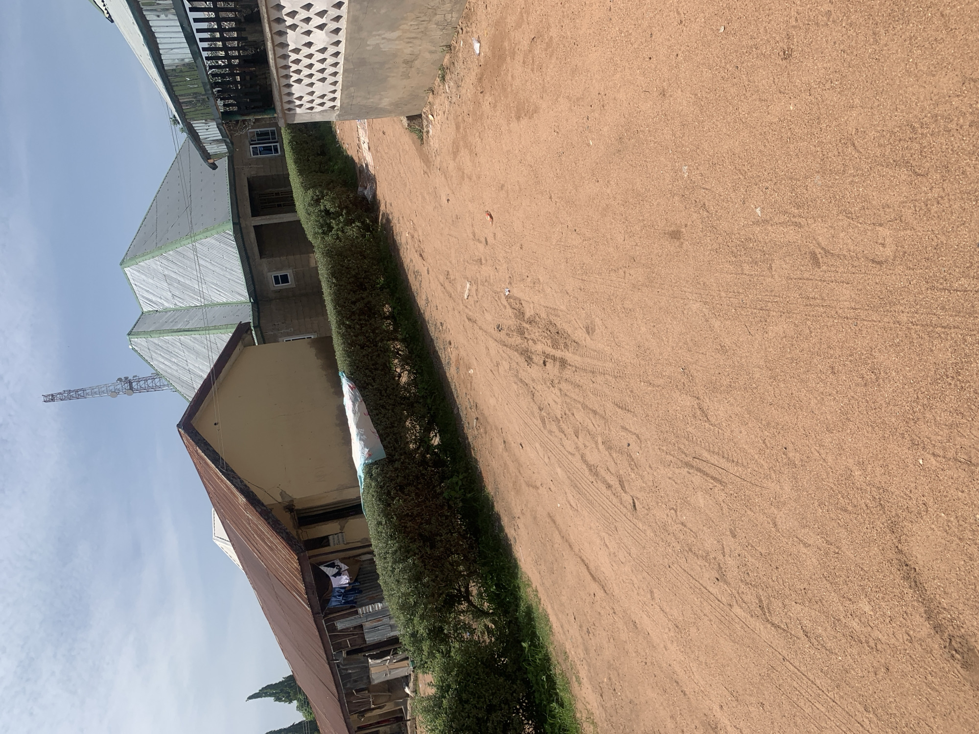 This is a ground-level, eye-level, medium shot of a dirt road in Jauro Yinu, Nigeria. The road is dusty and reddish-brown, with tire tracks visible. To the left of the road, there is a row of buildings with corrugated iron roofs and light yellow walls. Some laundry is hanging from lines in front of these buildings. A hedge separates the road from these buildings. Further back, a modern-looking building with a silver, multi-faceted roof is visible. To the right of the road, there is a low, patterned concrete wall, behind which is another building with a fence. A tall communication tower is visible in the distance. The sky is light blue with scattered clouds. The overall impression is one of a rural or semi-rural settlement.