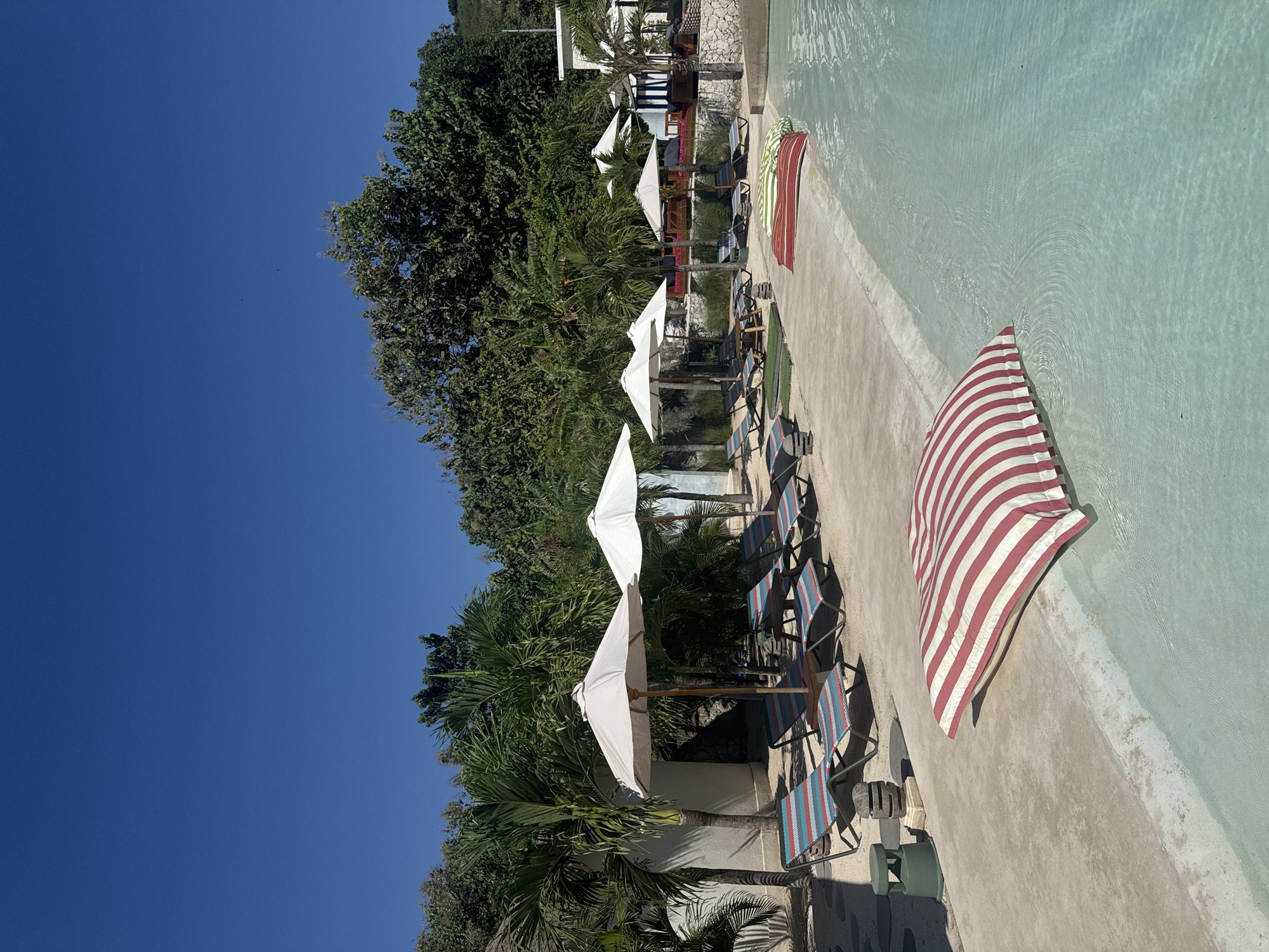 This image depicts a resort in Nusa Penida, Indonesia. The scene is an outdoor leisure area under a clear, deep blue sky, featuring a swimming pool, lounge facilities, and tropical vegetation.
In the background, a hillside covered with dense green foliage extends across the upper portion of the frame. The sky above is cloudless, with two small, dark, indiscernible specks visible in the upper left.
The midground showcases the resort's amenities. Several white-walled buildings with dark openings are partially visible amidst lush tropical plants, including numerous palm trees. A row of large, light-colored sun umbrellas with wooden poles provides shade. Beneath these umbrellas, multiple sun loungers with striped fabric (red, white, blue, and green) are arranged on a light-colored ground surface, which appears to be a mix of sand and concrete. A small, dark table is positioned between two of these loungers. Stone retaining walls are also present.
The foreground is dominated by a swimming pool on the right and a concrete deck on the left. The pool contains light turquoise water with gentle ripples. A large, rectangular beanbag or cushion with prominent red and white stripes rests partially submerged in the pool, extending onto the concrete deck. On the concrete deck to the left, a dark green cylindrical speaker and a small stack of irregular, natural-colored stones are visible. No individuals are present in the image. The scene is brightly illuminated by direct sunlight, casting distinct shadows.
