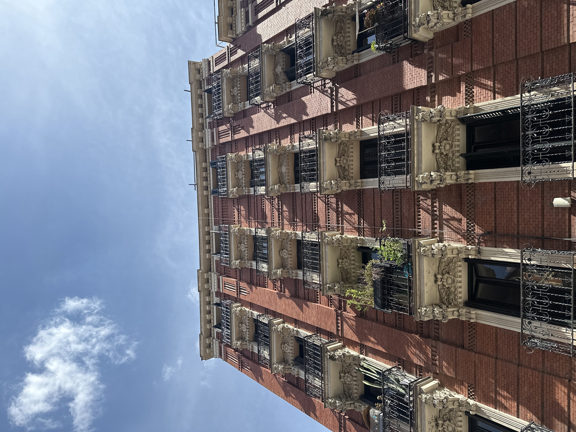 An upward-angled shot captures the ornate facade of a multi-story building against a clear blue sky with scattered white clouds. The structure, likely an apartment or residential building in Madrid, Spain, features a striking red brick exterior. Numerous windows are visible across several floors, each adorned with elaborate light-colored stone carvings and intricate black wrought-iron grilles or small balconies.
The architectural style suggests a historic urban setting. Many of the window frames are decorative, featuring classical motifs and volutes. Some windows have dark frames or shutters, and on a few balconies, potted plants or greenery are visible, adding a touch of life to the stone and iron. Strong, diagonal shadows are cast across the brickwork by the projecting architectural elements and window structures, indicating bright daylight, likely midday sun. No people are visible, and there is no discernible activity taking place. No text is visible on the building. The image emphasizes the detailed craftsmanship and grandeur of the architecture set against a serene sky.