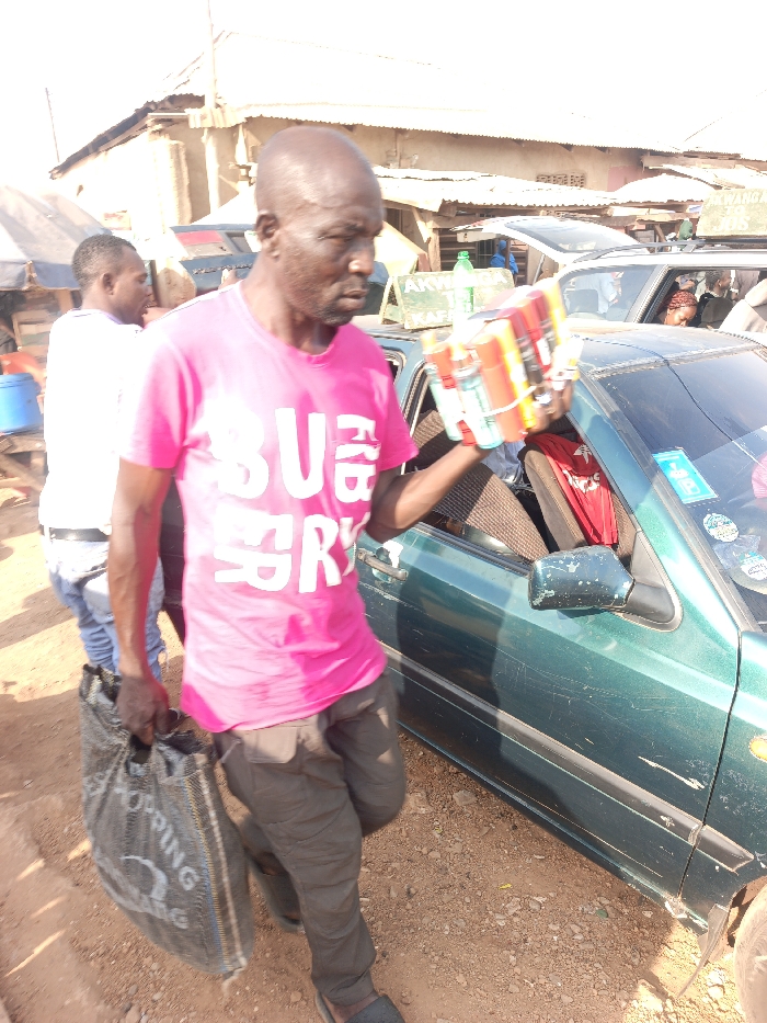 The scene depicts an outdoor, unpaved area in Akwanga, Nigeria, characterized by reddish-brown dirt and gravel ground. A prominent green and white sign in the background clearly displays "AKWANGA" and "KAFA."
In the foreground, an adult male, bald and with dark skin, walks from left to right. He wears a pink t-shirt with white block lettering, partially reading "BU BRRY," dark trousers, and dark sandals. In his extended right hand, he holds a bundle of approximately six aerosol cans, multi-colored and secured with elastic bands. His left hand carries a dark grey/black reusable shopping bag with white text including "SHOPPING" and "KWANG." He glances slightly to his right.
To the right of this individual, a dark green sedan is parked, its passenger side visible and the front passenger door ajar. The vehicle exhibits scratches on its bodywork and multiple stickers on the front passenger window. A red garment or item is visible on the passenger seat.
In the mid-ground, behind the primary subject, another male individual wearing a white t-shirt and light-colored jeans is partially visible, facing away from the camera. Further back, a white vehicle, possibly an SUV, with an open roof rack is parked. The background also features several low-rise structures with corrugated metal roofs, consistent with market stalls or simple buildings. A blue bucket or container is visible on the far left. The scene is illuminated by bright, natural daylight.