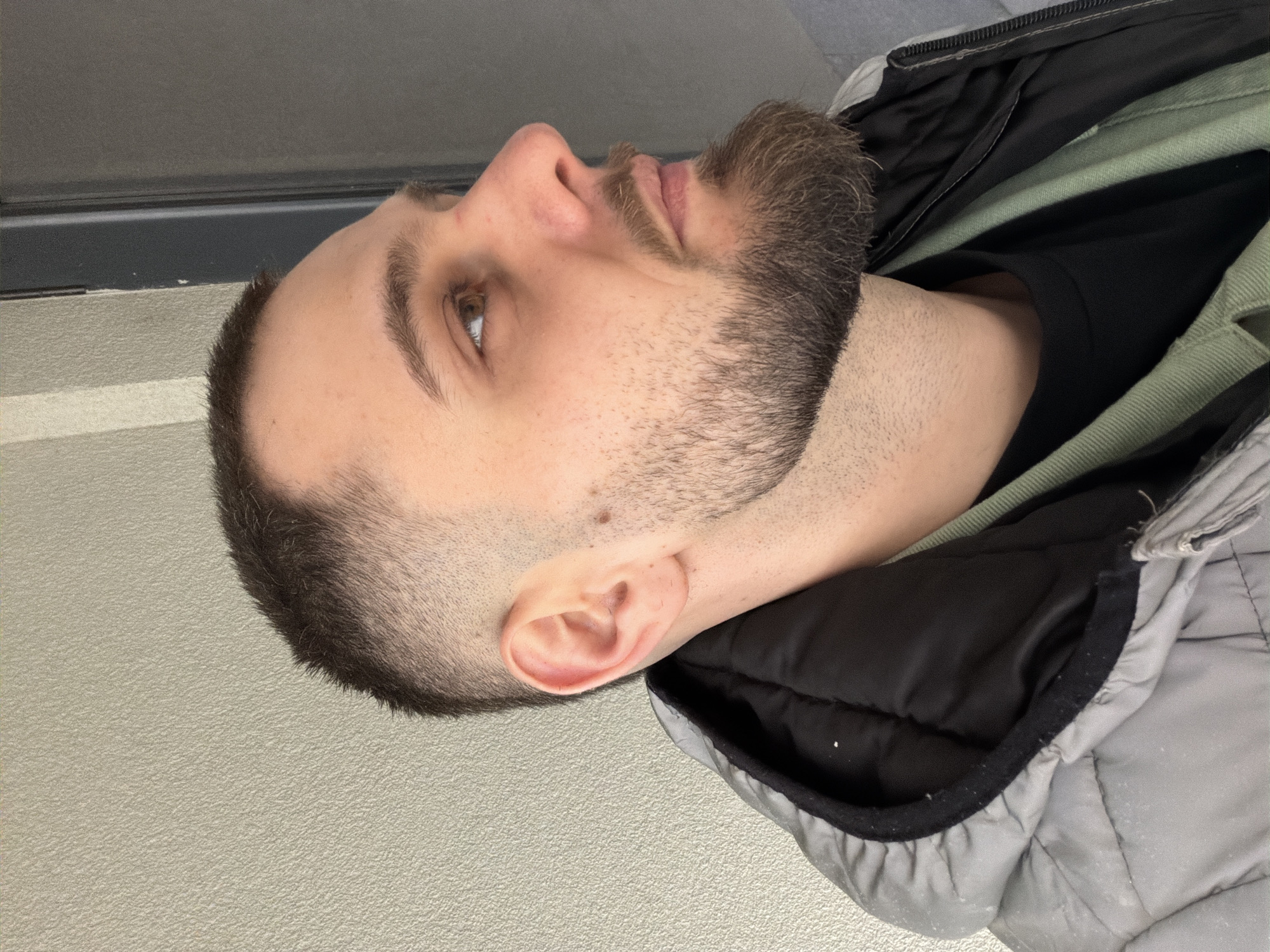 The image is a close-up, side-profile view of a light-skinned man. He has short, dark brown hair that is faded on the sides. He has a brown beard that connects with his hair. He has brown eyes. He is wearing a black t-shirt under a light green shirt, which is under a grey, quilted jacket with a black interior lining. The background is a plain, light grey wall. The scene is not recognizably related to Plovdiv, Bulgaria, as there are no distinguishing features or elements associated with that location.