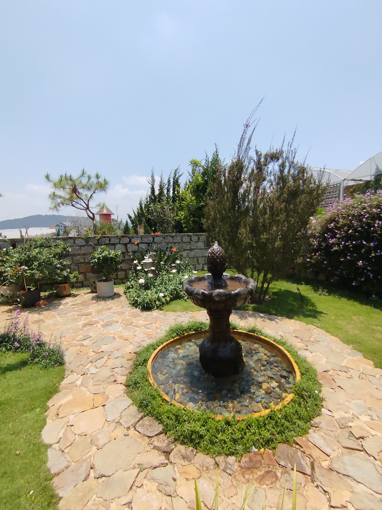 The image shows a stone fountain in a garden setting in Lang Biang - Đà Lạt, Vietnam. The fountain has a multi-tiered structure topped with a pineapple-shaped ornament. It sits in a circular basin filled with water and small rocks, encircled by green vegetation. The ground is paved with irregularly shaped flagstones. Behind the fountain are various plants and shrubs, including some blooming purple flowers, backed by a stone wall. In the background, trees and distant buildings are visible under a clear blue sky.