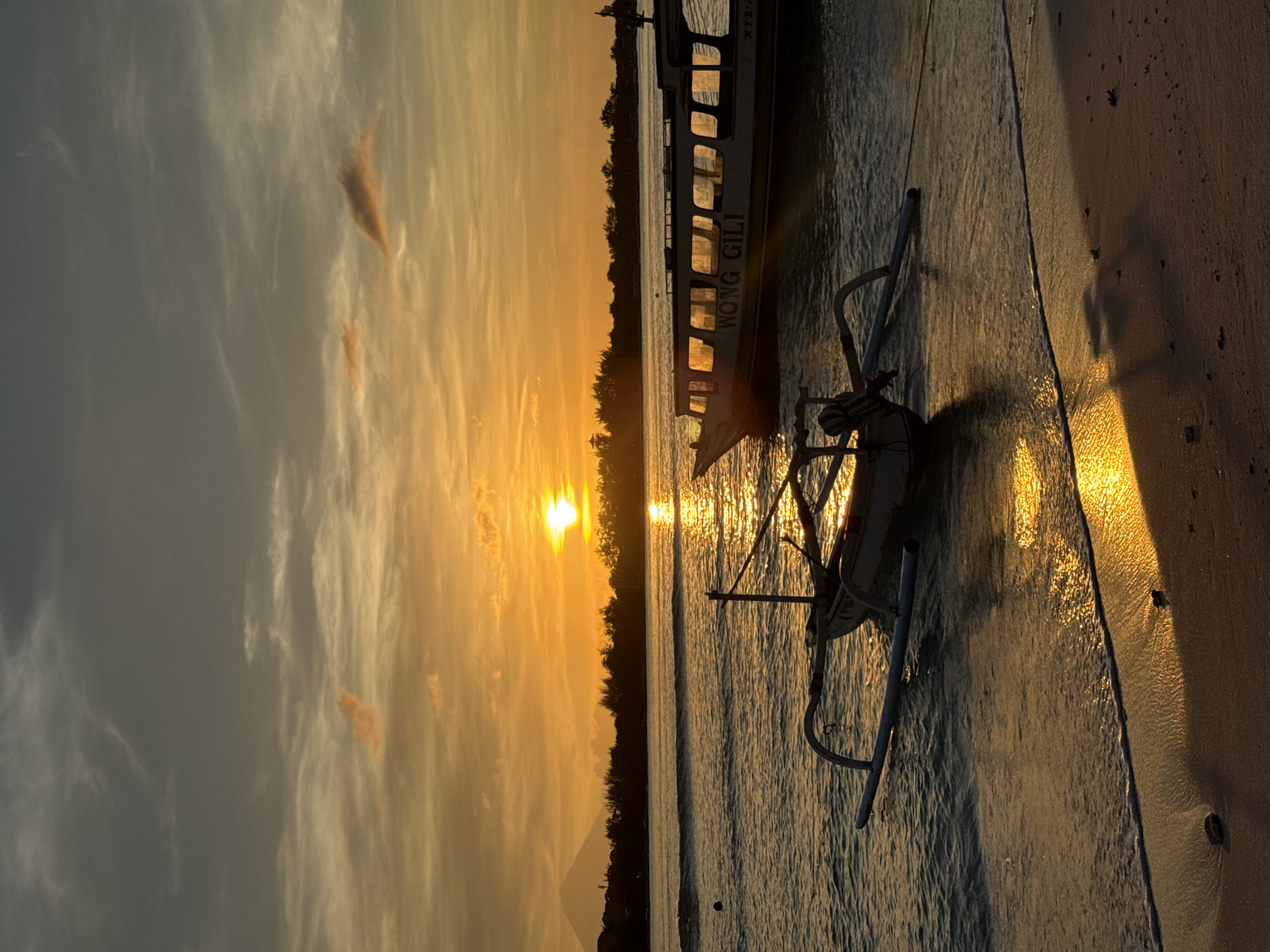 The image depicts a sunset over the ocean in an unknown location in Indonesia. A traditional outrigger boat is pulled up onto the beach. In the background, a larger boat is visible, bearing the words "WONG GILI" and "NTB". The sun casts a golden glow on the water, beach, and sky. Trees line the coastline. The sky is filled with clouds.