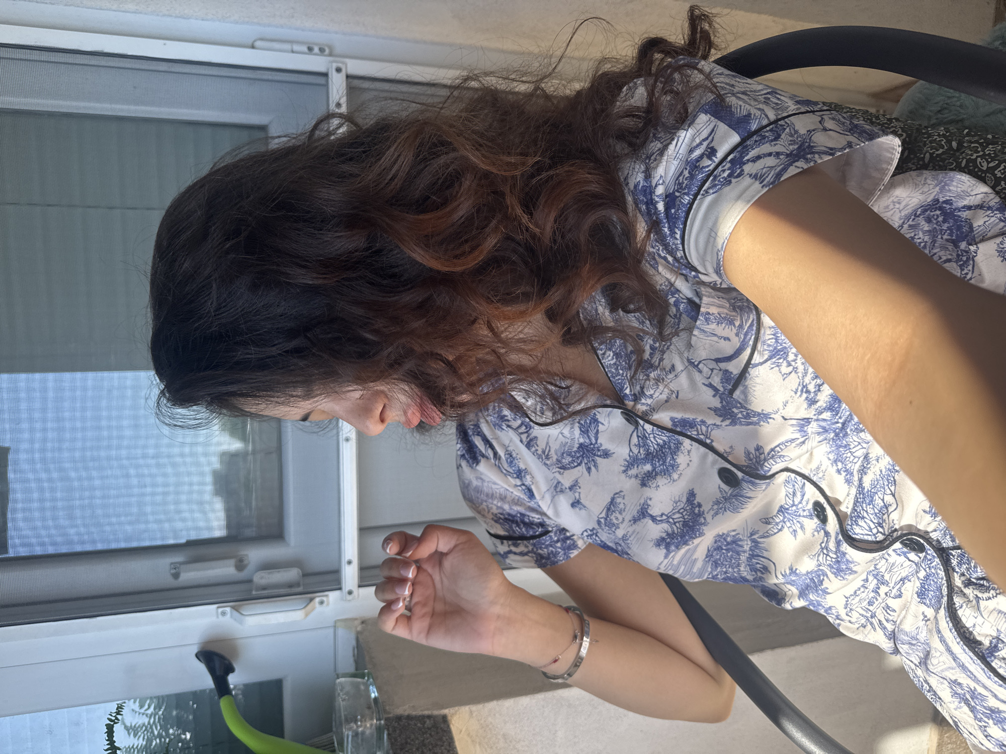 A woman with long, wavy brown hair, some with reddish highlights, is seated in a black chair, facing away from the camera and looking to her right. She is wearing a pajama top with a blue and white toile pattern. Her left hand is raised, with fingers curled inward. She is wearing a silver bracelet on her left wrist. The background features a window with a screen, a wall, and sunlight casting shadows. A green hose-like object is visible on the left side of the window. The scene appears to be on a balcony or near a window on an upper floor, possibly in an urban setting like Plovdiv, Bulgaria.