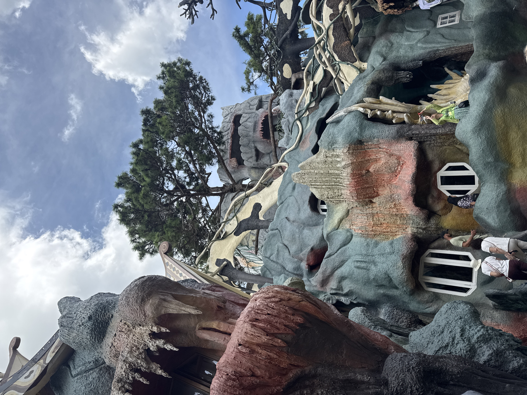 The image depicts the exterior of the Hang Nga Guesthouse, also known as the "Crazy House," in Da Lat, Vietnam. The architecture is organic and whimsical, resembling a fantastical tree or cave. The structure is made of concrete and painted in various earthy tones, with colorful accents. Several people are visible, appearing to be tourists exploring the unique building. One person in a green outfit is standing on a ledge, looking upwards and seemingly interacting with the structure. Another group of people in casual attire are walking along a pathway. The surrounding environment includes lush green trees and a cloudy blue sky. The building itself is characterized by irregular shapes, winding pathways, and unusual window designs.