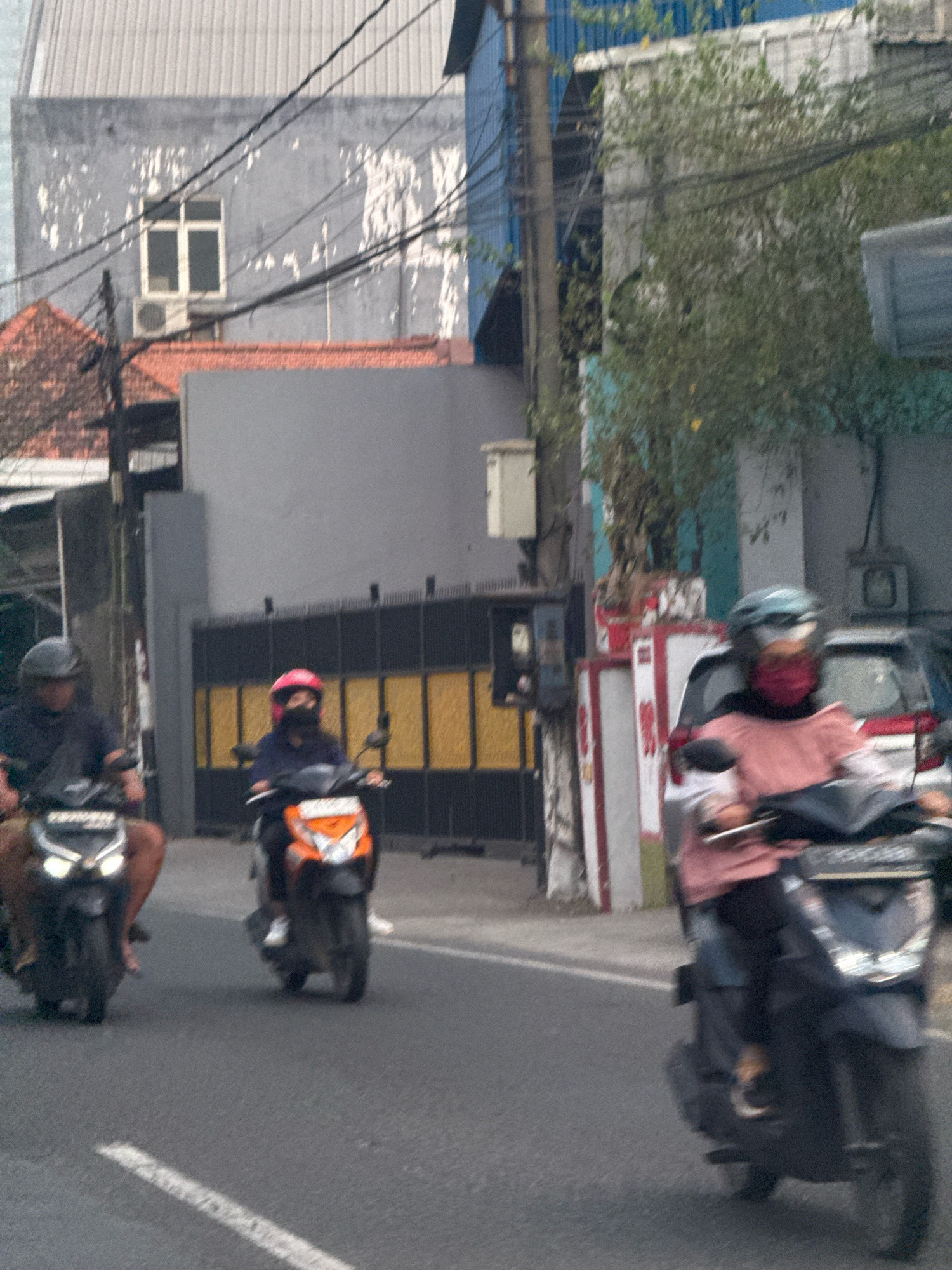The image depicts a street scene in Surabaya, Indonesia, with several individuals riding motorcycles. In the foreground, two motorcycles are visible from the side and slightly from the front. The motorcycle on the left has its headlights on, illuminating the road. The rider on this motorcycle is wearing a dark helmet and a dark top. The motorcycle on the right is orange and black, and its rider is wearing a pink helmet and a dark top, with a red mask covering their face.
Further back, another motorcycle is seen, with a rider wearing a blue top and a red helmet, also with a mask covering their face. Behind this motorcycle, parts of a car and other vehicles are visible. The environment consists of buildings with corrugated metal roofs and grey facades, along with power lines crisscrossing overhead. A large black and yellow striped gate is present in the background, suggesting the entrance to a property or compound. The road surface is asphalt, with a white line marking the lane. The overall impression is a busy urban street during daylight, with traffic moving along.