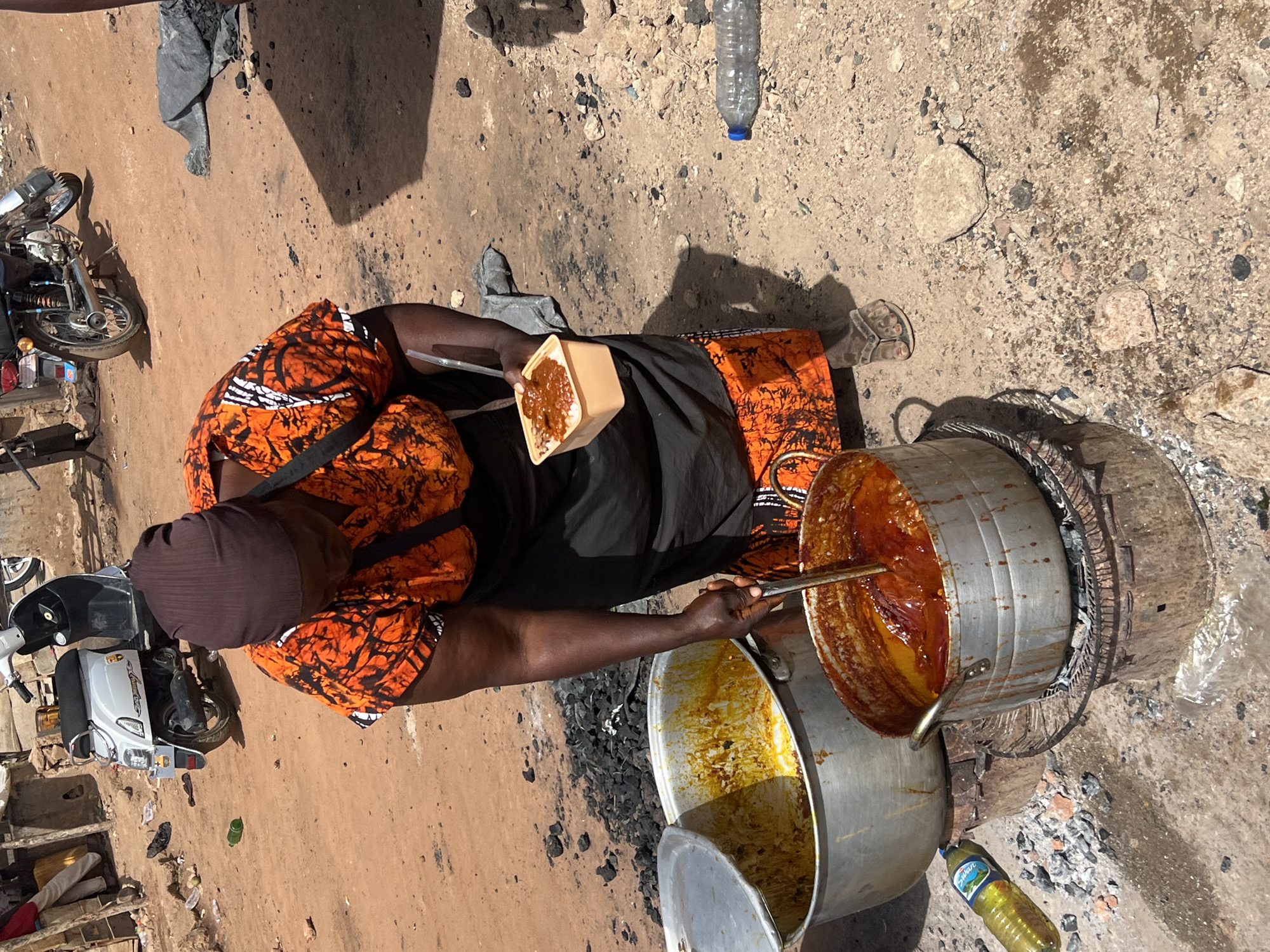 This outdoor scene in Jos, Nigeria, captures a woman engaged in street food vending under bright daylight, indicated by strong shadows. The main subject is a dark-skinned woman, seen from a high angle, wearing a vibrant orange and black patterned top, a dark skirt, and a dark head covering. She is actively cooking or serving food from large metal pots set on the dusty, unpaved ground.
She holds a long metal spoon, stirring a rich, reddish-orange stew or sauce in one large aluminum pot. This pot rests on a circular, makeshift stove structure, likely fueled by charcoal, as evidenced by the dark ash and charcoal pieces scattered around it. Beside this main pot is another large aluminum pot, partially open, which appears to contain a lighter, possibly yellow, dish or ingredient. In her left hand, she holds a light brown rectangular container filled with food, showing a base layer of what looks like rice topped with the same reddish stew, suggesting she is preparing a meal for a customer.
The immediate surroundings feature a rough, light brown ground, strewn with small stones and some debris. Various objects are visible: a clear plastic water bottle and a green plastic bottle with the brand "Sultan" (likely cooking oil) lie on the ground near the cooking area. In the background, to the left, a silver motorcycle and a white scooter are parked, further indicating an urban or semi-urban street setting. The scene reflects a common method of street food preparation and sale in the region.