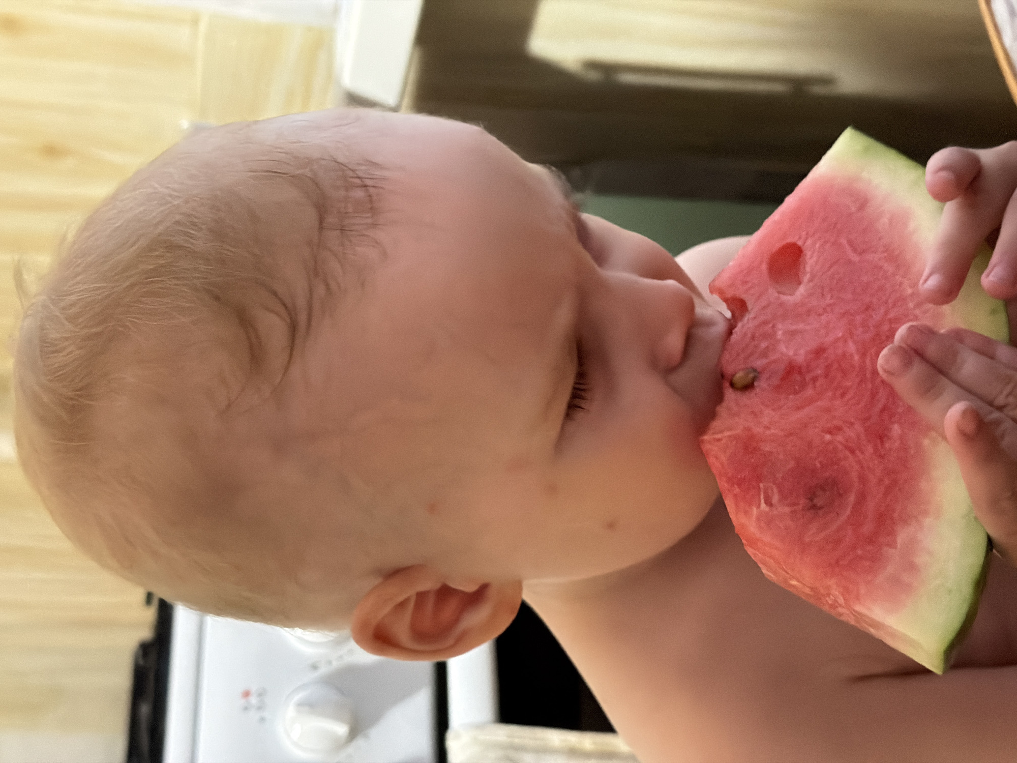 A close-up shot shows a baby, possibly a toddler, eating a slice of watermelon. The watermelon is held in the baby's hands and has some seeds visible. The location is not determinable from the image.