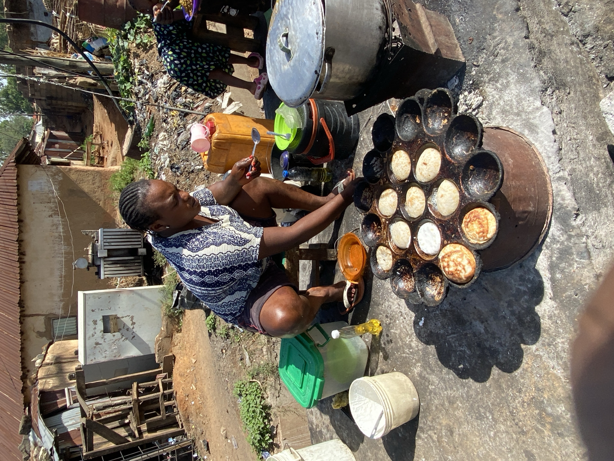 The image captures a woman in Bukuru, Nigeria, preparing food outdoors. She sits next to a large, circular, multi-cavity pan on a metal stand, with what appear to be small cakes or pastries cooking in the cavities. A metal pot sits nearby. She is using a spoon, presumably to add ingredients or manipulate the cooking items. Various containers, including plastic buckets and jugs, are scattered around her. Behind her is a building and some discarded debris. The scene is outdoors, with evidence of a partially constructed or dilapidated structure in the background.