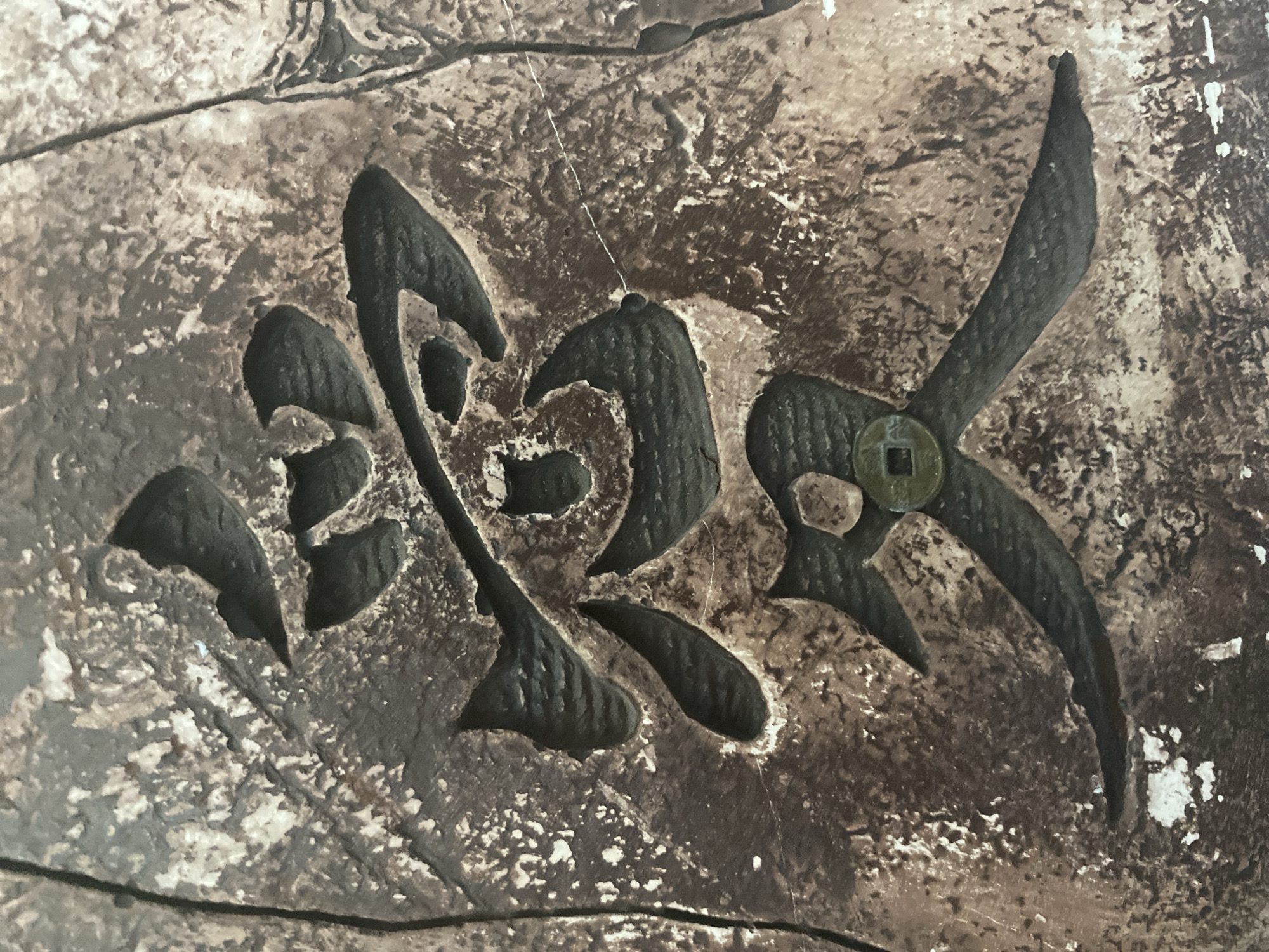The image displays a close-up, overhead view of a textured, weathered surface with a carved inscription. The inscription is a single, complex character, possibly of East Asian origin, rendered in a dark, recessed style against the lighter, rough background. The surface appears to be made of stone or ceramic, with patches of white and lighter brown coloration interspersed with darker brown and black areas, suggesting age and exposure. A small, round coin with a square hole in the center is embedded within the lower portion of the carved character, adding a unique detail. The lighting is even, highlighting the texture of the material and the depth of the carving. There is no visible indication of the city of Kemerovo, Russia, or any elements that would place the image in that specific location. The scene is static and focuses solely on the artifact itself.