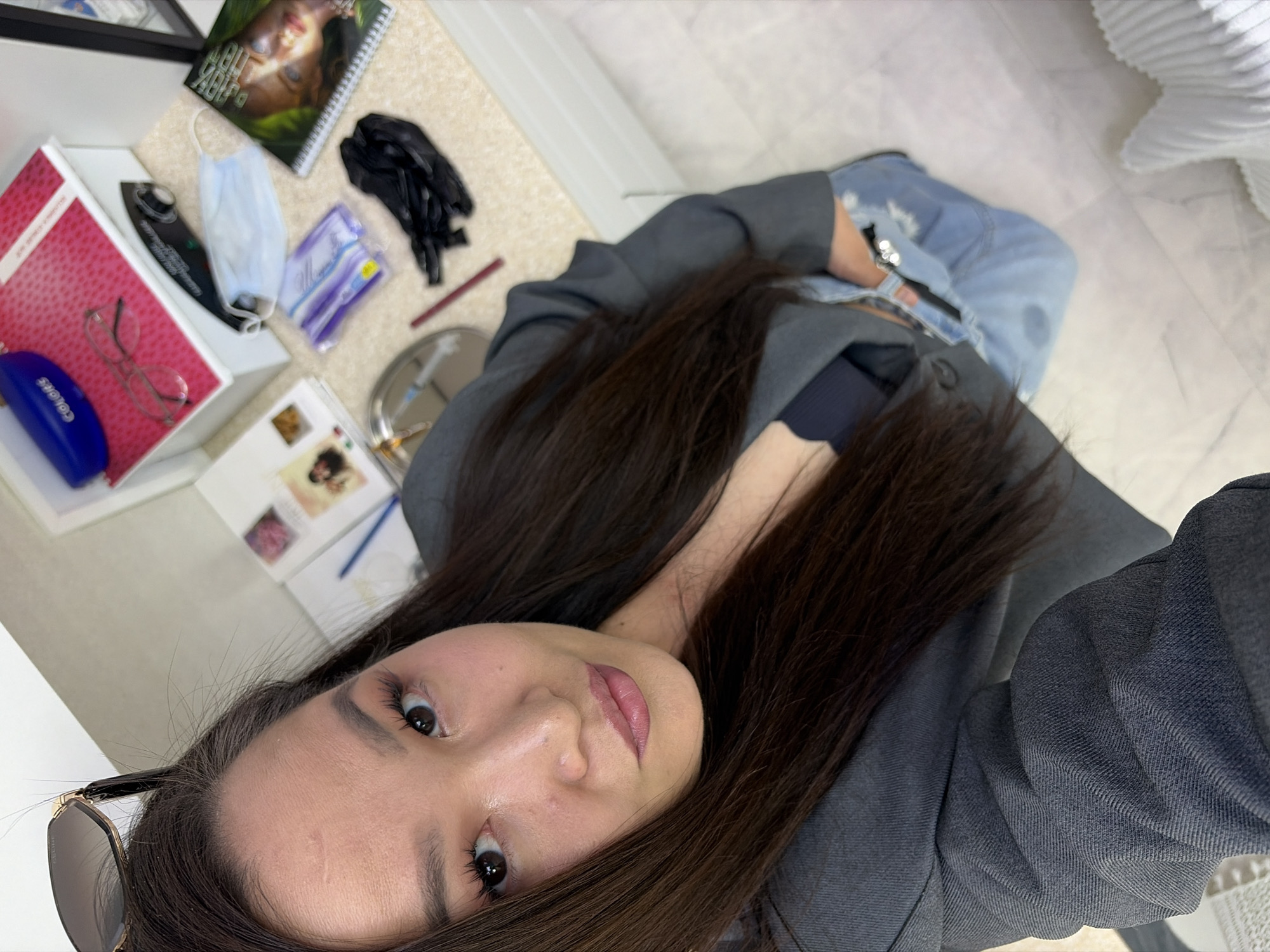 The image is a selfie taken indoors. The subject is a woman with dark hair, wearing a dark-colored top and a gray blazer. Sunglasses are tucked into her hair near her forehead.
The background shows a desk or table with various items, including a calendar or notebook, a face mask, black gloves, and packages of wet wipes. There's also a box with glasses, a blue case, and a small silver tray. The surface under the desk appears to be light-colored tiled flooring. The location is not identifiable as being in any specific city.