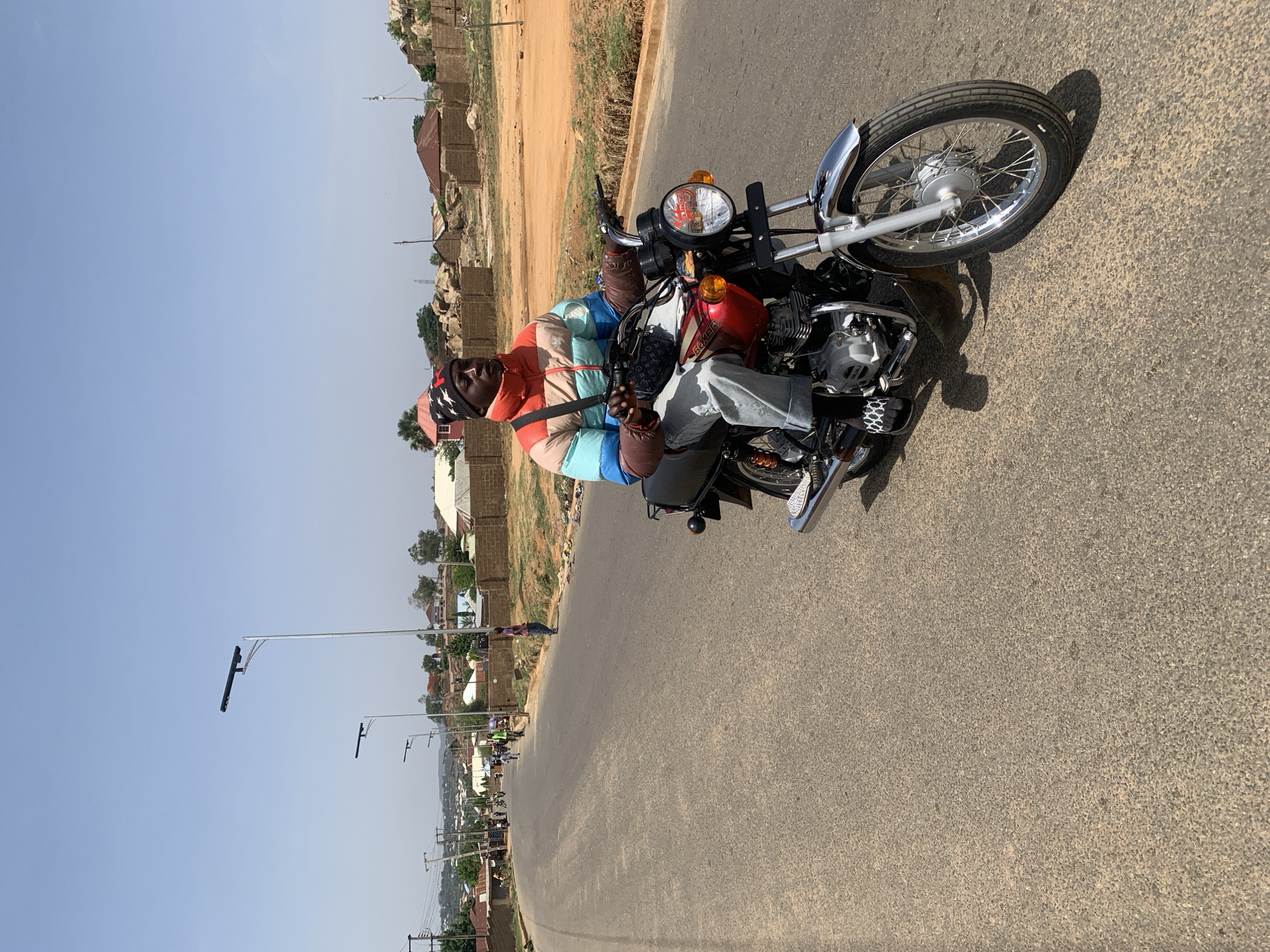 A man rides a red and black motorcycle on a paved road in Gura Top, Nigeria. He is wearing a jacket, jeans, and a black cap with white stars. The motorcycle has a chrome front fender and a silver front wheel. The road is lined with buildings and trees. The sky is blue and clear. There are street lights on the side of the road. There are other people and motorcycles in the distance. The terrain shows some buildings set on a hill.