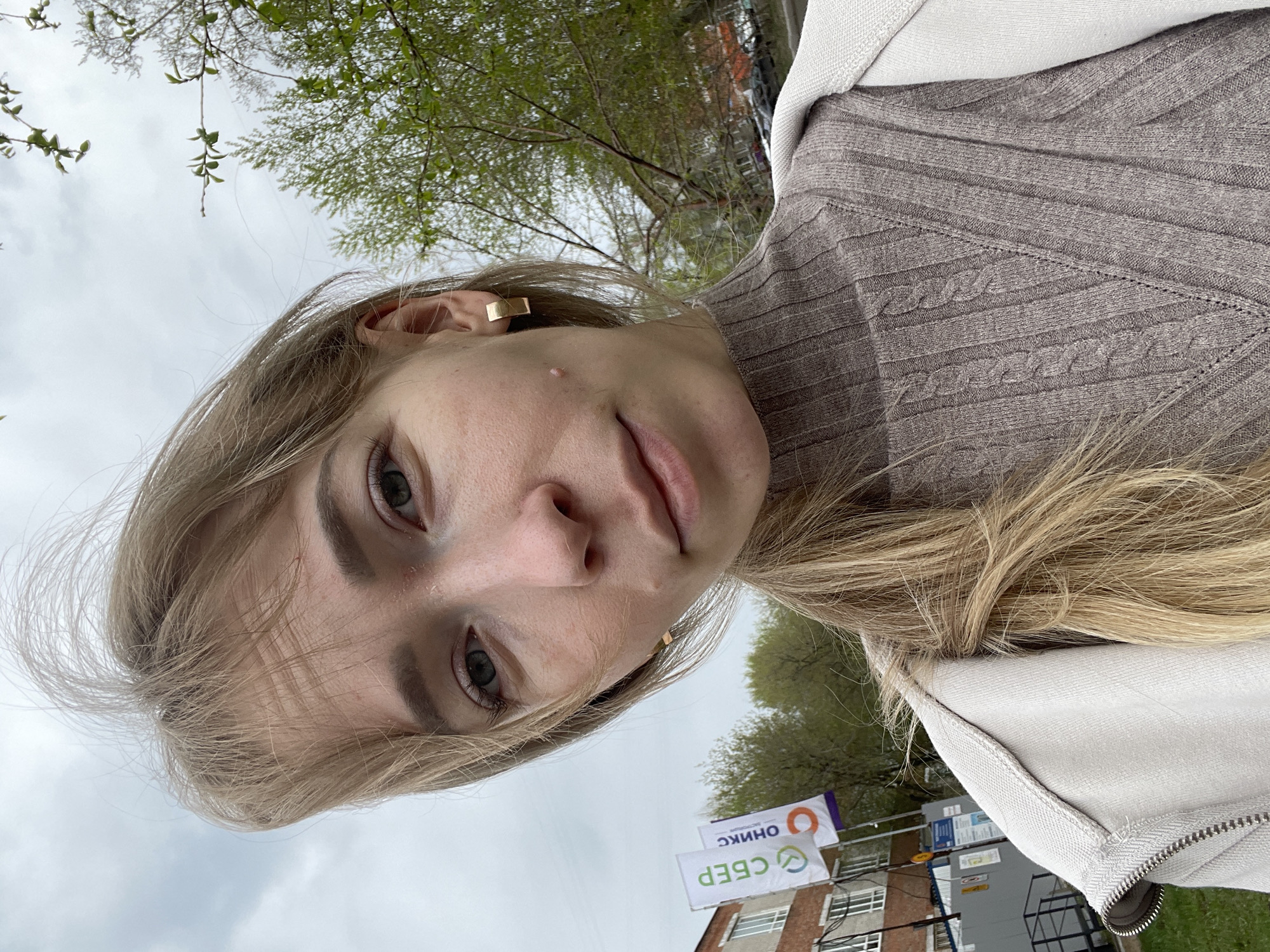 A selfie-style photograph shows a young woman in an outdoor setting. Her face is the primary focus, capturing her features including blonde hair, eyes, and a square earring. She is wearing a beige turtleneck sweater and a light-colored jacket. The background contains trees with green foliage and a cloudy sky. Buildings are visible in the background. Russian advertisements of the Сбербанк (Sberbank) and ОНИКС are featured. Based on the advertisements and architectural style of the buildings, the location is in Perm, Russia.