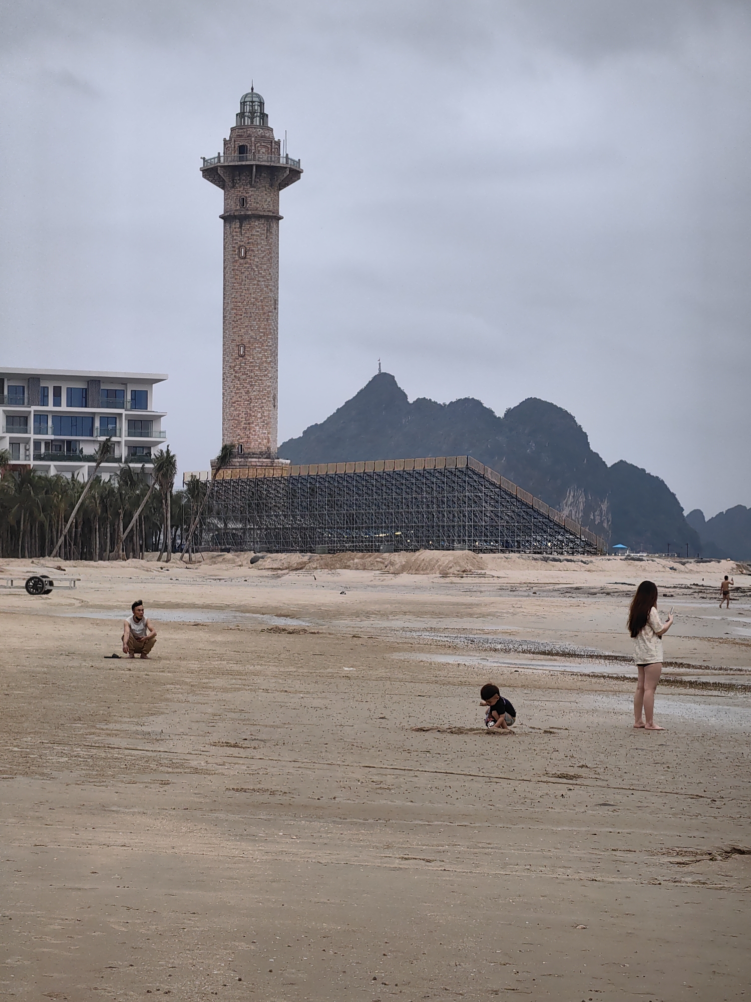 The image presents an overcast day at a beach in Bai Chay Ward, Vietnam, featuring a mix of natural elements and human development. The foreground consists of a wide, sandy beach with damp sections and subtle tidal patterns.
Several individuals are present on the beach. On the left, a man wearing a light tank top and pants is squatting down, observing the scene. Towards the center-right, a young child in a dark top is kneeling and playing in the sand. On the far right, a woman with long dark hair, dressed in a light-colored shirt and shorts, stands barefoot, engrossed in her phone. A distant figure is also visible walking on the beach further to the right.
Prominently featured in the mid-ground is a tall, slender tower, resembling a lighthouse, constructed from brick-like material and capped with an ornate, greenish lantern room. To its left stands a modern, multi-story white building, likely a hotel or resort, characterized by its clean lines and balconies. A cluster of palm trees stretches between this building and the beach. Behind the tower and extending across the middle of the image is a large, dark metal framework structure, possibly part of a construction project like a roller coaster or observation deck, built upon a sandy embankment.
The background is dominated by rugged, forested mountains, typical of karst landscapes, silhouetted against a uniformly gray, cloudy sky. The weather appears overcast, casting a soft, diffused light across the entire scene. No distinct text is visible in the image. The scene captures a moment of casual beach activity amidst significant coastal development.