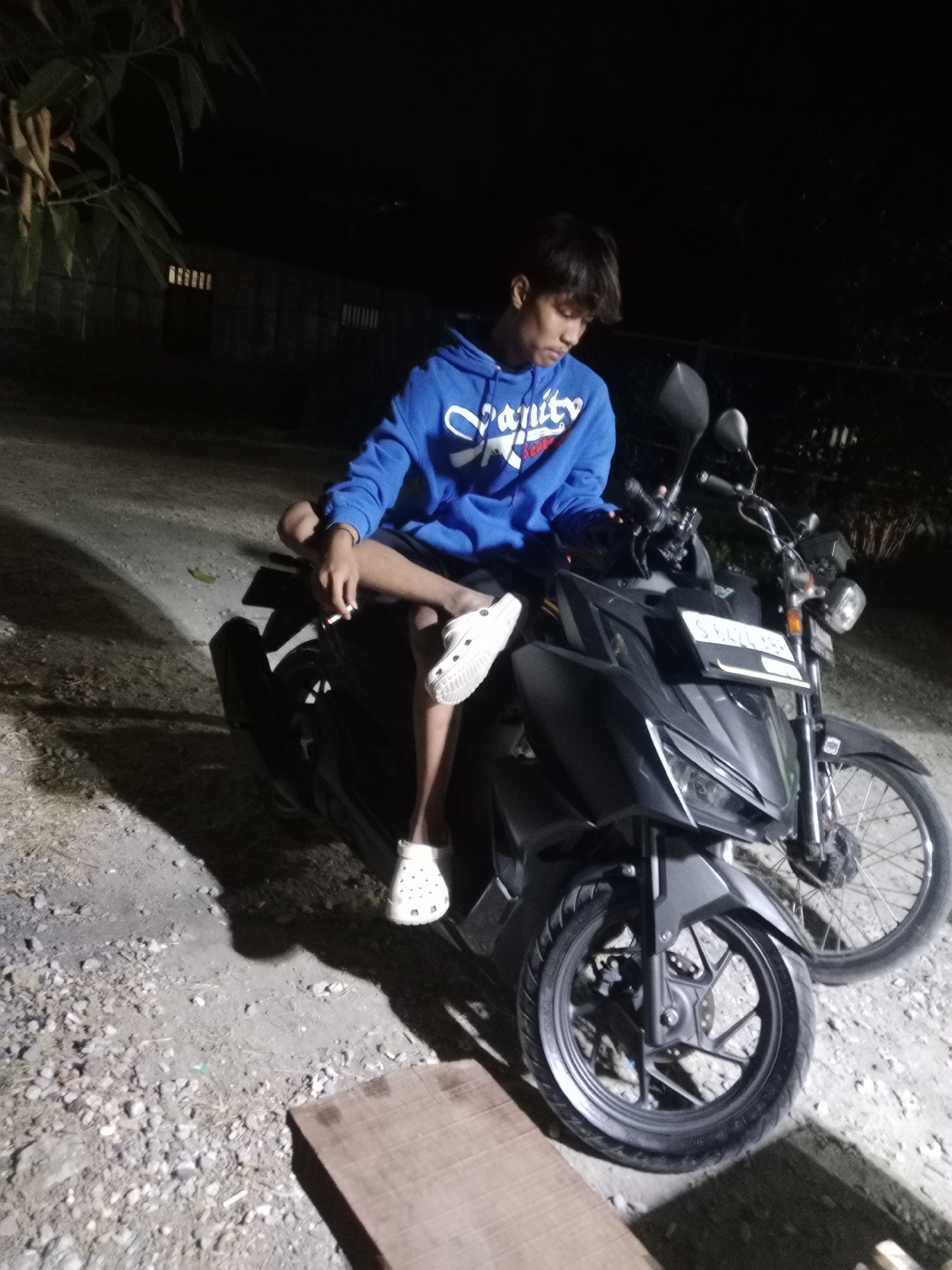 A young male sits on a black motorcycle at night, holding a lit cigarette in his right hand. He is wearing a blue hoodie with white lettering and dark shorts. His left leg is crossed over his right, and he is wearing white Crocs on both feet. The motorcycle has a license plate that reads "S 8476 ABB". The ground is covered in gravel and dirt. In the background, there is a wooden fence and some trees. The scene appears to be in Glagah Wangi, Indonesia.