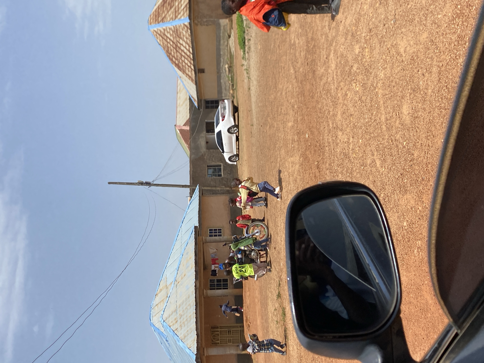 In Bar Jirgi Summa, Nigeria, on a sunny day, a group of children are gathered around a motorcycle in a dirt courtyard. A man in a blue cap and a neon yellow shirt stands by the motorcycle, which has a green frame and a green bag attached to the back. Several children, dressed in casual clothing, are looking at the motorcycle and interacting with each other. One child is holding a book, while another is carrying a yellow backpack. In the background, there are several houses with corrugated metal roofs, some painted blue. A white sedan is parked in front of one of the houses. A side-view mirror of a vehicle is in the foreground, reflecting the sky and the interior of the car. The ground is covered in reddish-brown dirt and small stones.