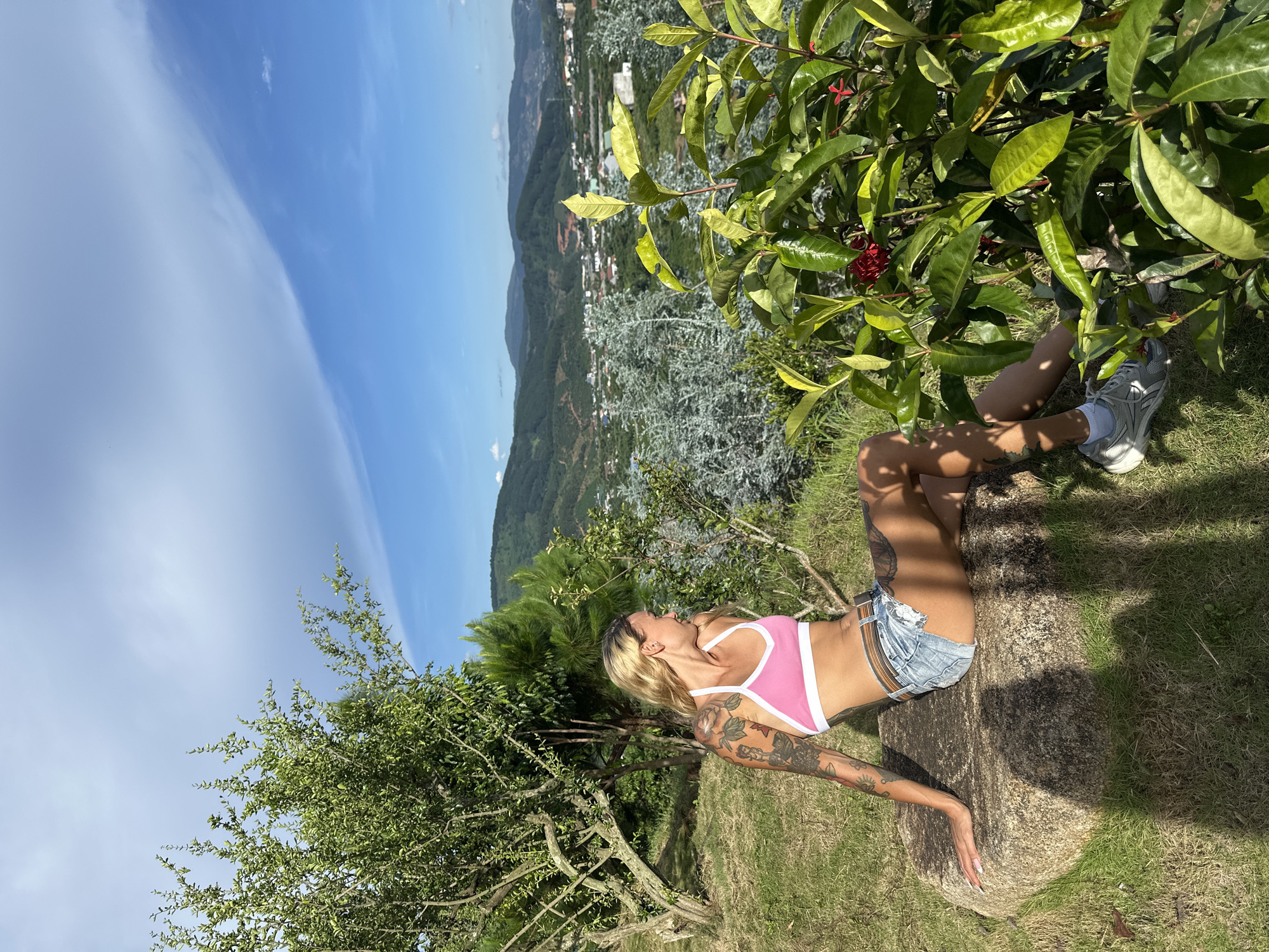 A woman with blonde hair and tattoos on her arms is lying on a large rock outdoors. She is wearing a pink and white sports bra and denim shorts. Behind her, a lush green bush with red flowers is visible. The background shows a panoramic view of a valley with rolling hills and a town nestled within. The sky is bright blue with some wispy clouds. The scene suggests a sunny day, likely in the late morning or early afternoon, given the strong shadows cast on the rock. The location appears to be a scenic overlook in Thôn 3, Vietnam, offering a stunning vista of the landscape. The woman's pose and expression convey a sense of relaxation and enjoyment of the natural surroundings.