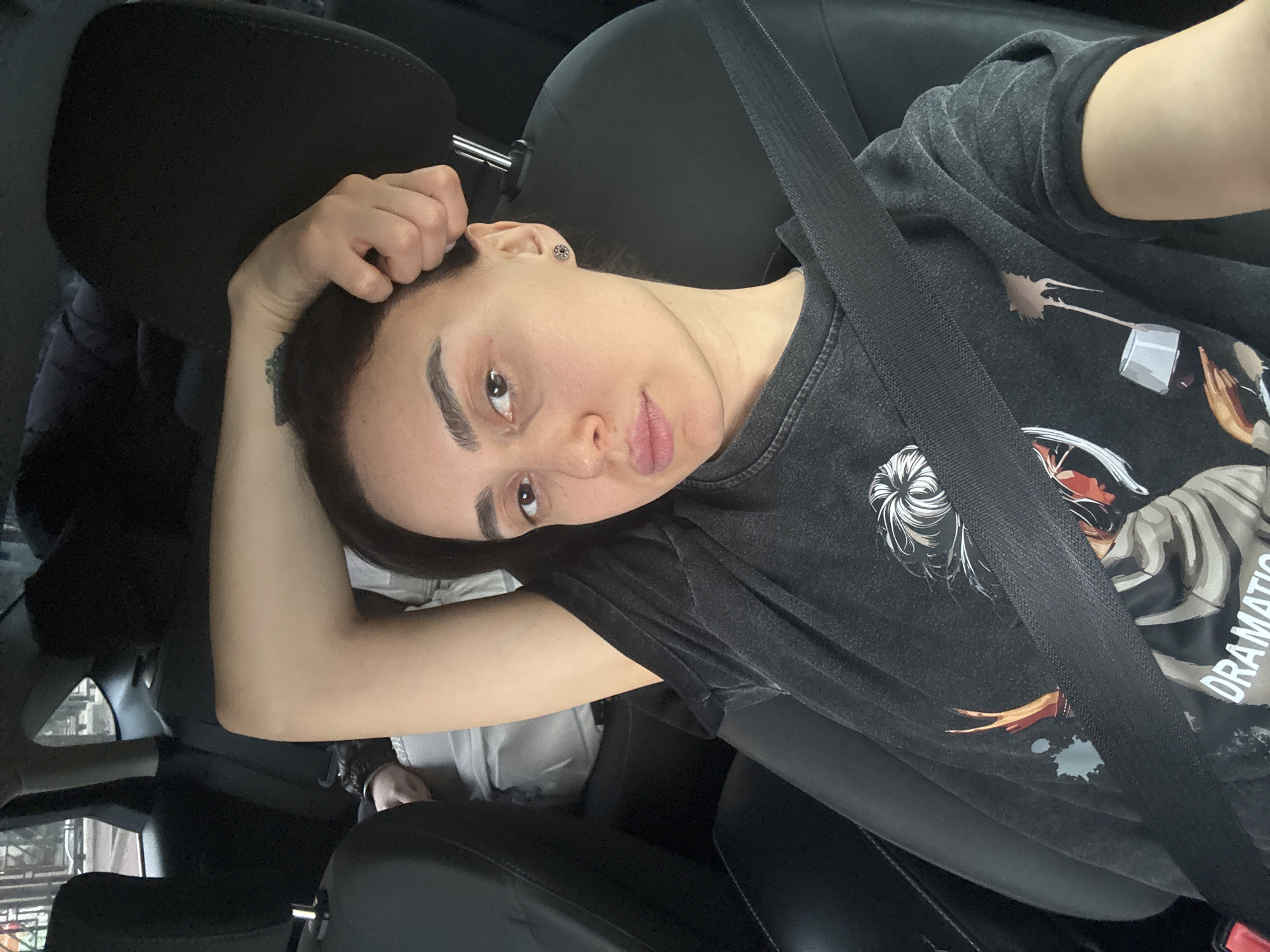 A selfie taken by a female passenger in a car. The passenger is wearing a black graphic t-shirt and a seatbelt. The car's interior is black. The location is unknown and there is no way to identify the city or country based on the visual information.