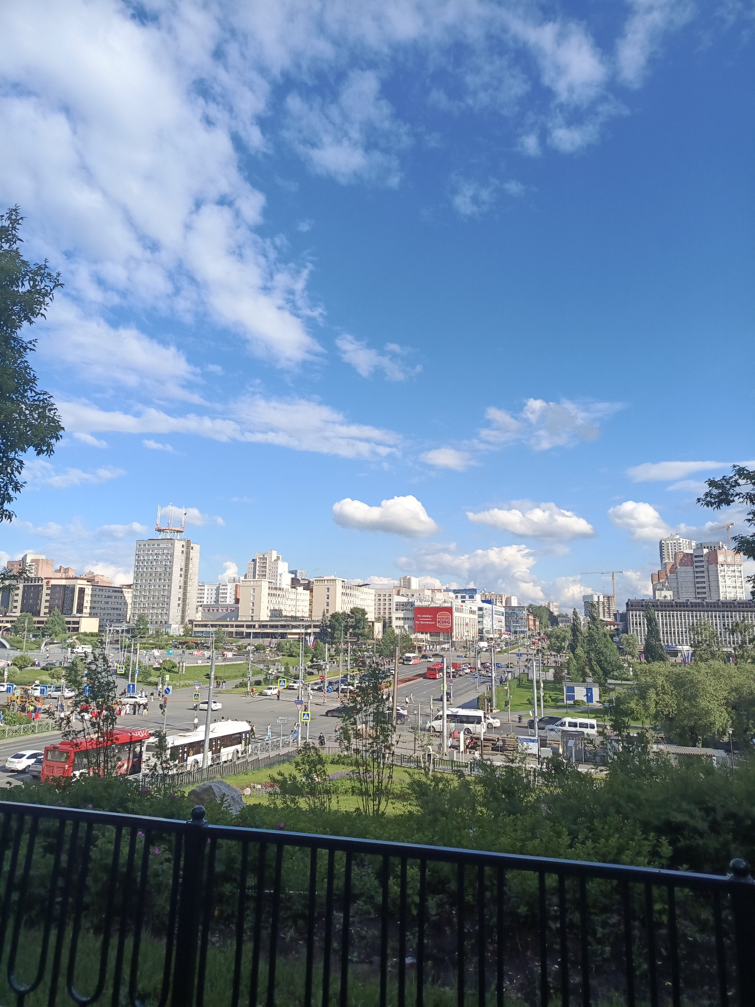 This is a high-angle, wide shot of the city of Perm, Russia, on a clear, sunny day. The sky is a vibrant blue with scattered white, puffy clouds. In the foreground, a black metal fence with vertical bars and rounded finials runs horizontally across the bottom of the frame, obscuring the lower portion of the scene. Beyond the fence, lush green foliage and trees are visible, with some pink flowers interspersed.
The middle ground depicts a busy city intersection with multiple lanes of traffic. Cars, buses, and vans are driving on the road. A prominent red bus and a white trolleybus are stopped near a pedestrian crossing. People are visible walking on the sidewalks and crossing the street. Streetlights and traffic signals are present, indicating a well-developed urban infrastructure. Several pedestrian crossings with yellow signage are visible, and one has a traffic light.
In the background, a panorama of city buildings stretches across the horizon. The architecture varies, with some buildings appearing to be older, while others are modern high-rises. One tall, grey building in the center of the frame is topped with a distinctive structure resembling a ship or a sail. Other buildings have multiple stories and exhibit a mix of architectural styles. Green trees and parks are interspersed among the buildings, adding a touch of nature to the urban landscape. Cranes suggest ongoing construction in some areas. Several large billboards, one with red lettering, are visible on the side of a building. The overall impression is a dynamic and active city center under a bright summer sky.