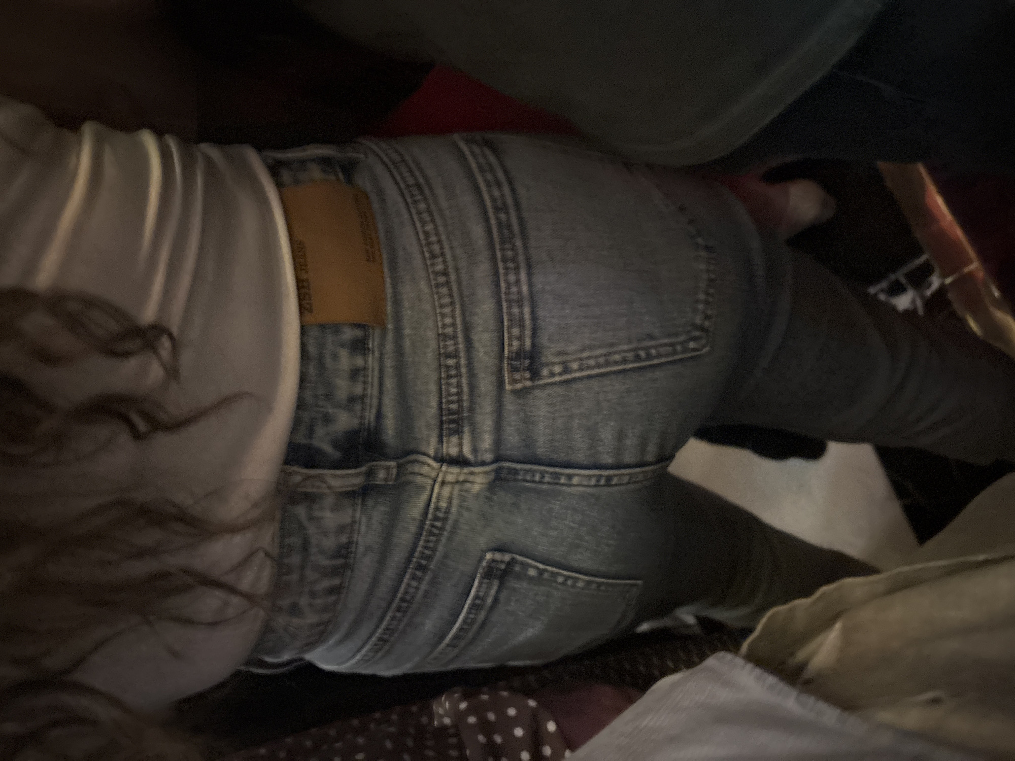 The image shows a close-up view of the lower back and legs of a person wearing light-wash denim jeans and a white ribbed top. The jeans have a leather patch on the back with embossed text that appears to read "ZARA JEANS". The scene is dimly lit, with some of the surroundings out of focus and appearing dark. The person is standing, and their back is to the camera. The environment suggests an indoor setting, possibly a public space given the presence of other people partially visible on the sides of the frame, one wearing a dark green garment. The overall impression is that of a candid, casual snapshot. There is no specific information in the image to indicate the location of Estosadok, Russia.