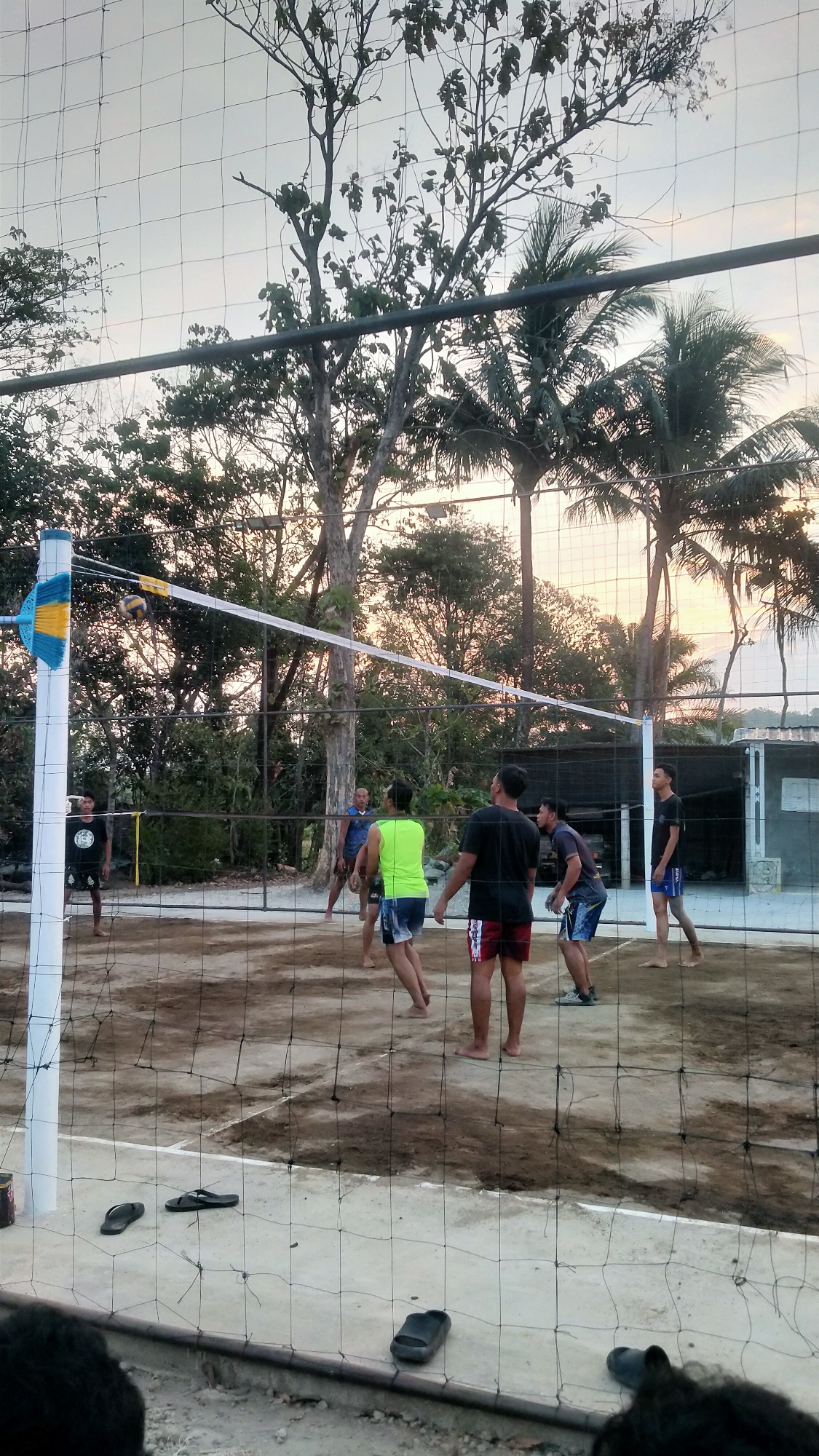 This is a medium shot, eye-level, outdoor photograph taken at a volleyball court in Ketoyan, Indonesia, during sunset. The sky displays a pale orange hue, indicating the sun is low on the horizon. The court is made of sand and dirt, with a net strung across its width. Several men are playing volleyball. In the foreground, a net with a grid pattern is visible. On the court, from left to right, a man wearing a black t-shirt and black shorts stands on the far side. Next to him, a man in a neon green tank top and blue and white patterned shorts is positioned. To his right, a man in a black t-shirt and red patterned shorts is facing away from the camera. Further back, another man in a grey t-shirt and blue patterned shorts is bending down. On the right side of the court, a man in a black t-shirt and blue shorts stands facing towards the net. Tall trees with lush foliage and palm trees are scattered around the perimeter of the court. A simple building with a grey roof and white walls is visible in the background to the right. Several pairs of flip-flops are scattered on the ground near the net in the foreground.