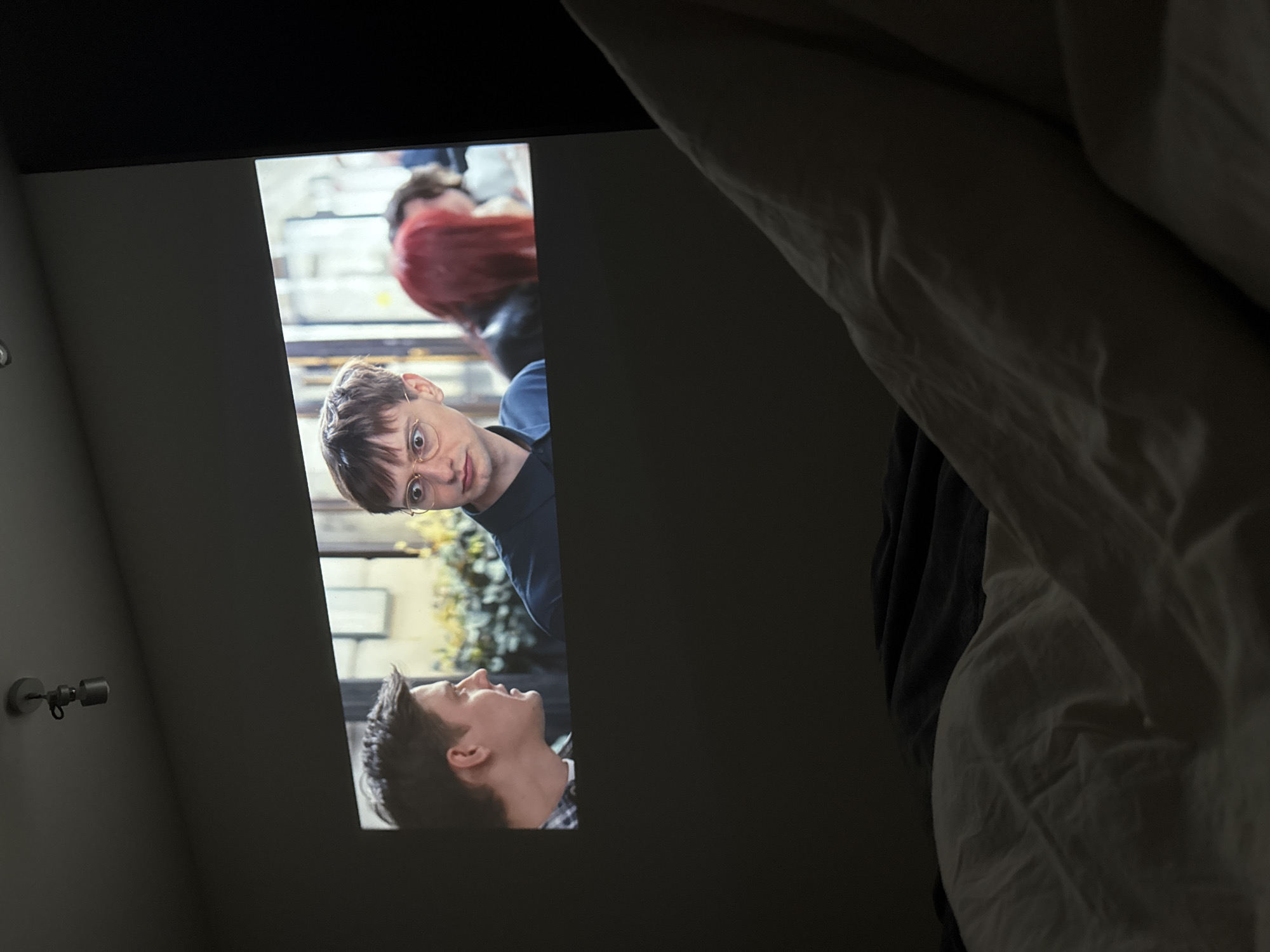 The image appears to show a movie or TV show projected onto a wall. Two male characters are visible, one wearing glasses and looking directly at the viewer, while the other is looking up. The background of the projected image seems to be an outdoor setting with greenery. To the left of the projected image is a wall-mounted light fixture. To the right of the projected image is a bed, with a white comforter/duvet. The city and location are indiscernible.