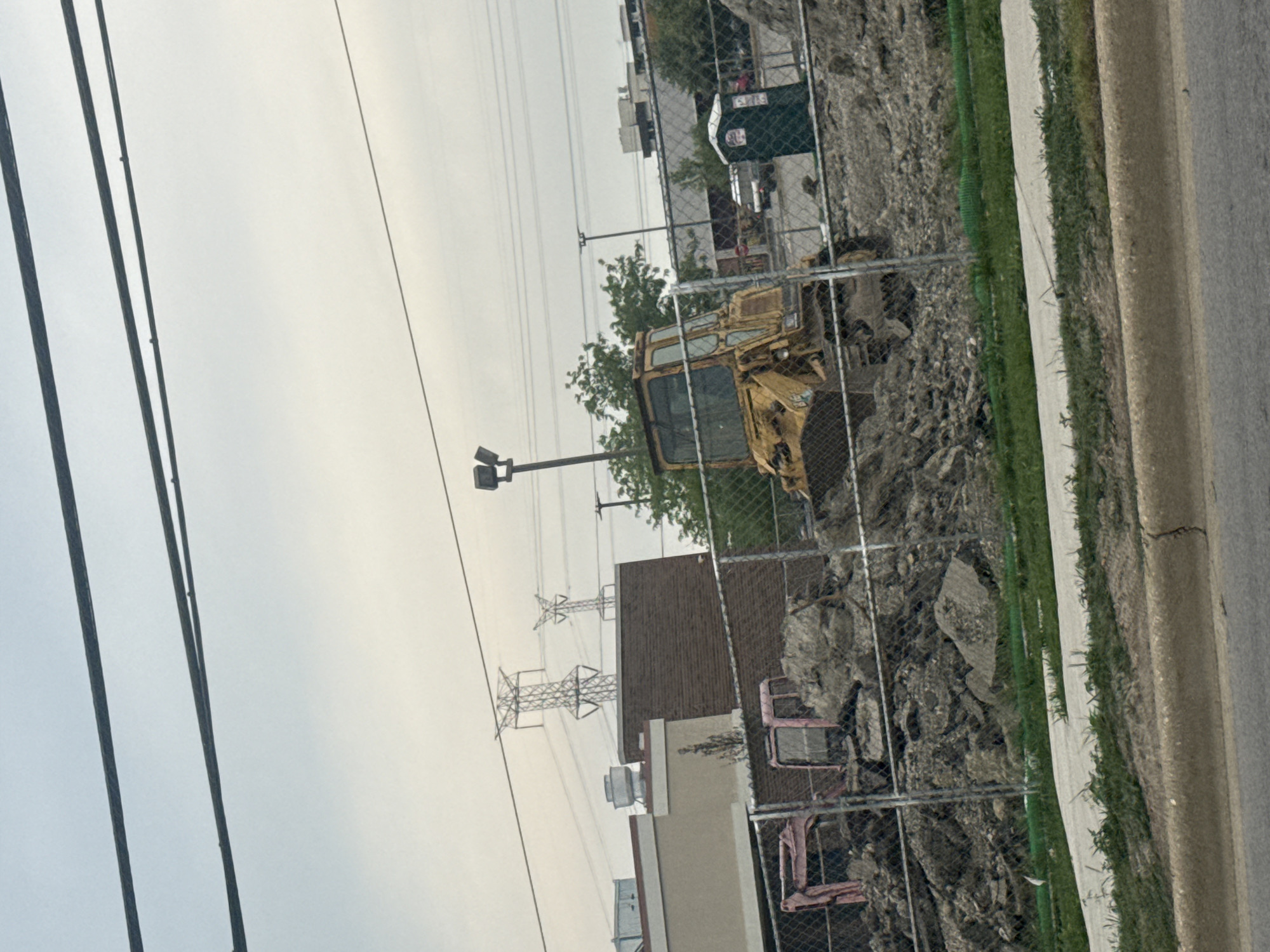 The image depicts a construction site in Niles, Illinois, USA. Two heavy machinery vehicles, a yellow bulldozer and a pink excavator, are present. The bulldozer is positioned in the center right of the frame, partially obscured by debris. The pink excavator is located to the left, also amidst piles of dirt and rubble. A green portable toilet is situated to the right of the bulldozer. In the background, several large electrical towers are visible against an overcast sky, and buildings suggest an urban or suburban setting. A chain-link fence runs across the foreground, separating the construction area from the street. The ground in front of the fence is covered with grass and a concrete path.