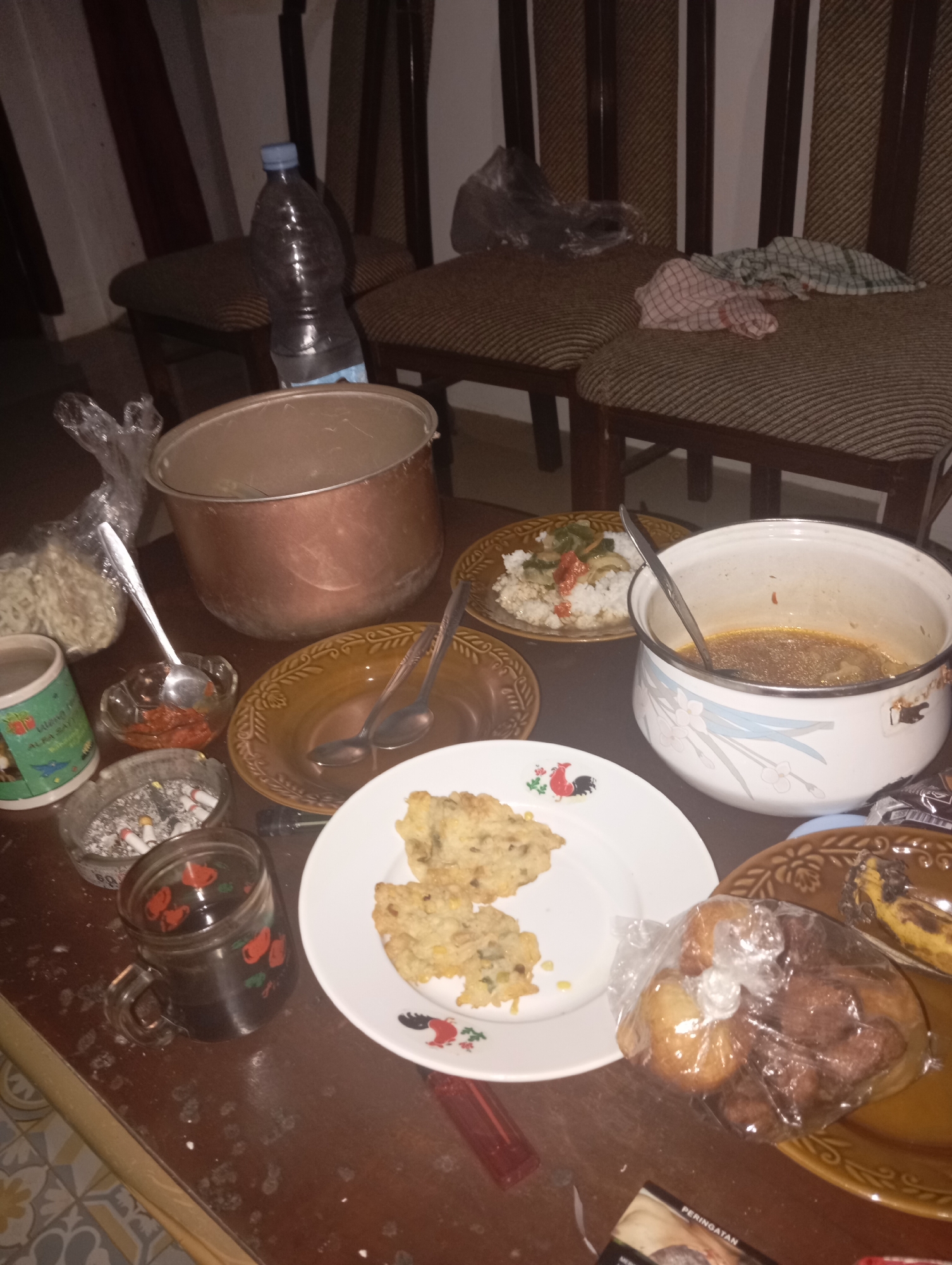 The image depicts a dining table in what appears to be a residential setting in Kedungbenda, Indonesia. The table is covered in various food items and dining utensils, suggesting a meal in progress or recently concluded.
On the table, there is a large, copper-colored pot, likely used for cooking. Next to it, a white pot with a floral design contains a reddish-brown liquid, possibly a soup or sauce. A plate of white rice with some green vegetables and red chili peppers is also visible. Two smaller brown ceramic plates are present, one with two spoons and the other empty, ready for use.
In the foreground, a white plate holds two pieces of fried food, possibly fritters or tempura, garnished with a rooster design. To the left, a glass mug with a red floral pattern is filled with a dark liquid, likely coffee or tea. An ashtray filled with cigarette butts and a lighter rests nearby. A small glass bowl contains a red chili paste or sauce. A plastic bag contains several pieces of fried dough or pastries.
Other objects on the table include a bottle of water, a plastic bag containing what appears to be crackers or chips, and a book of matches or a cigarette pack with Indonesian text. Two chairs with striped upholstery are visible in the background. The overall lighting suggests an indoor setting, possibly at night. The scene conveys a sense of informal dining and consumption.